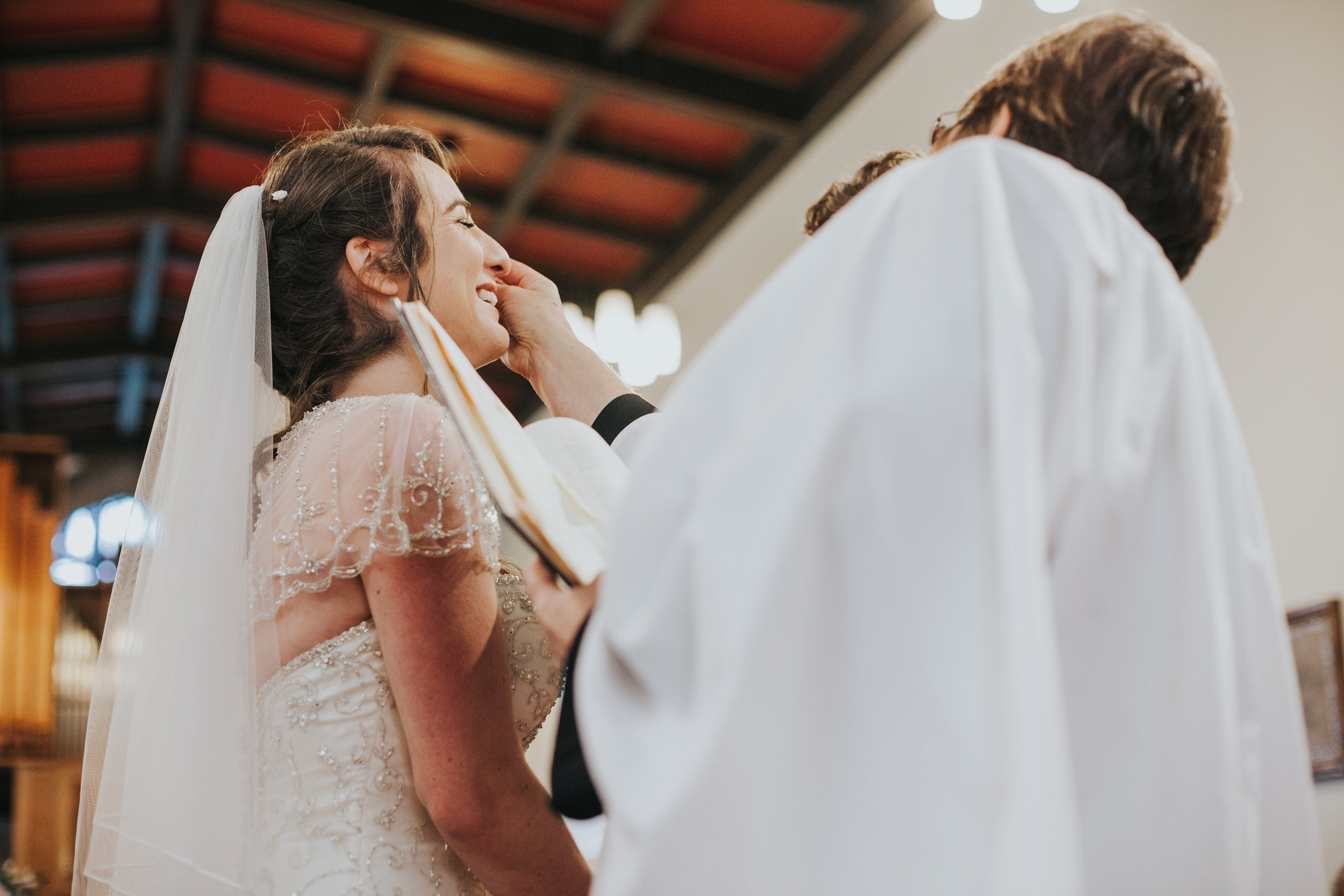  Female vicar wipes tears from brides face, while bride laughs. 