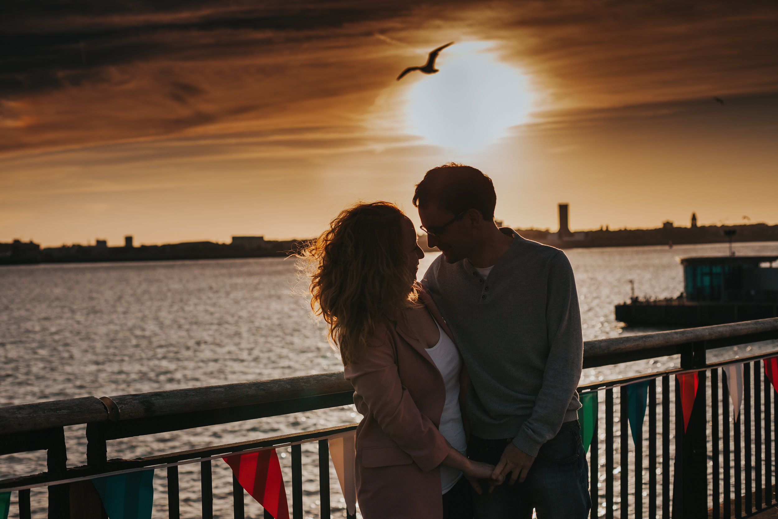 Couple come together in front of sun set, a single bird in the sky behind them just over the sun. 