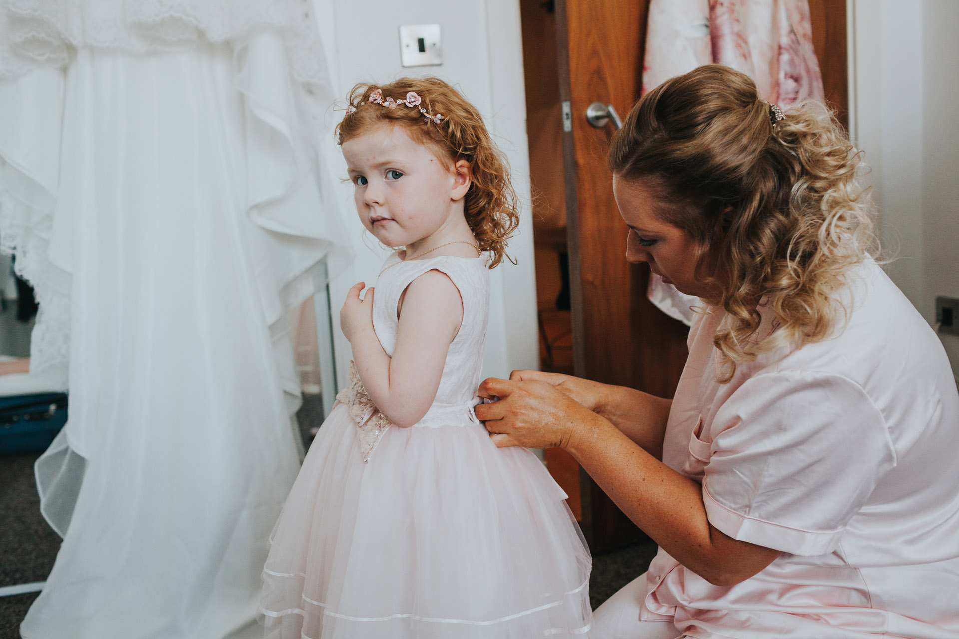 Flower girl had her buttons done up by bridesmaid, the brides wedding dress hangs in the background.