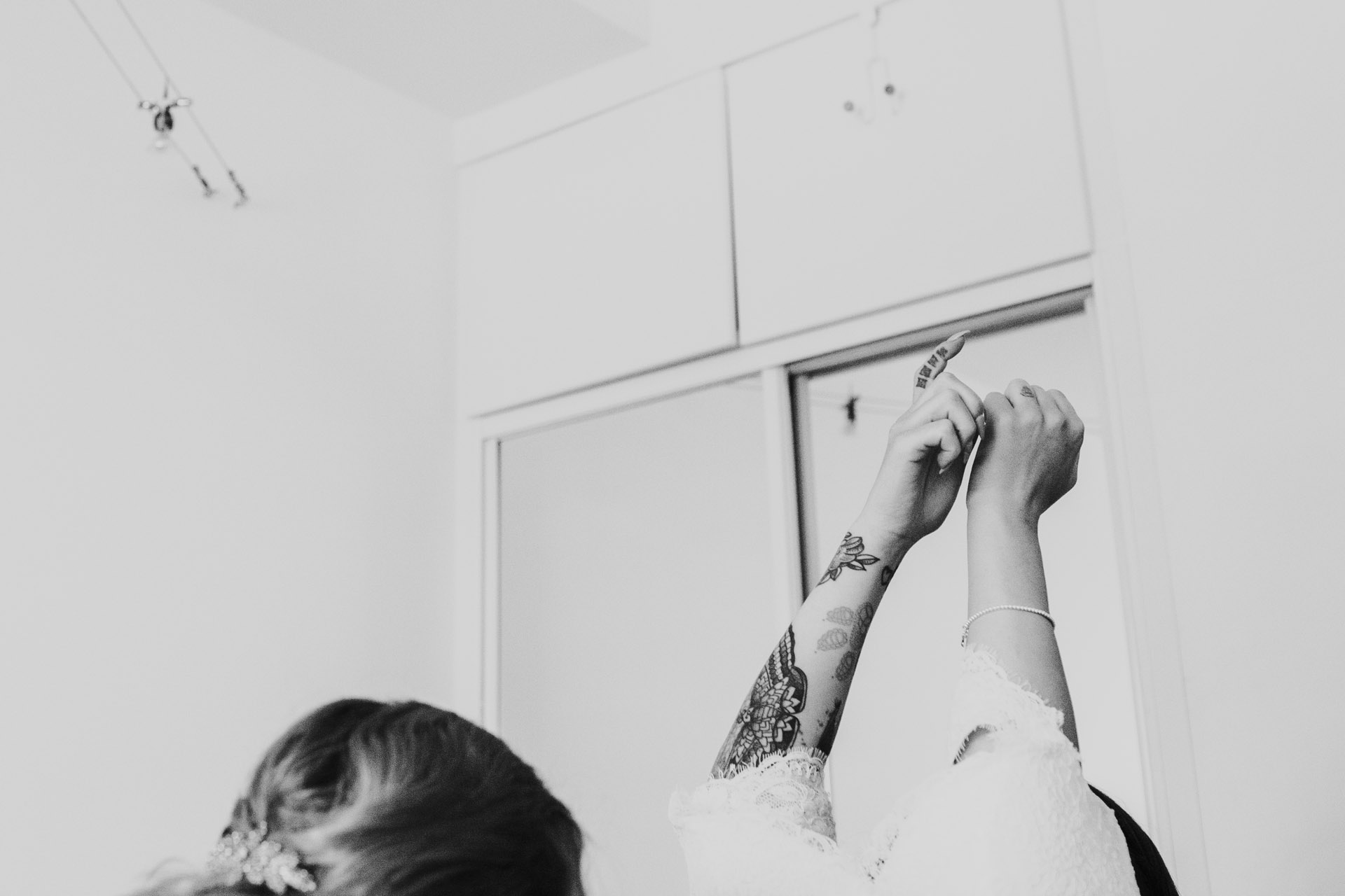 Brides tattooed arm reaches up as she puts on her wedding dress. Image processed in black and white