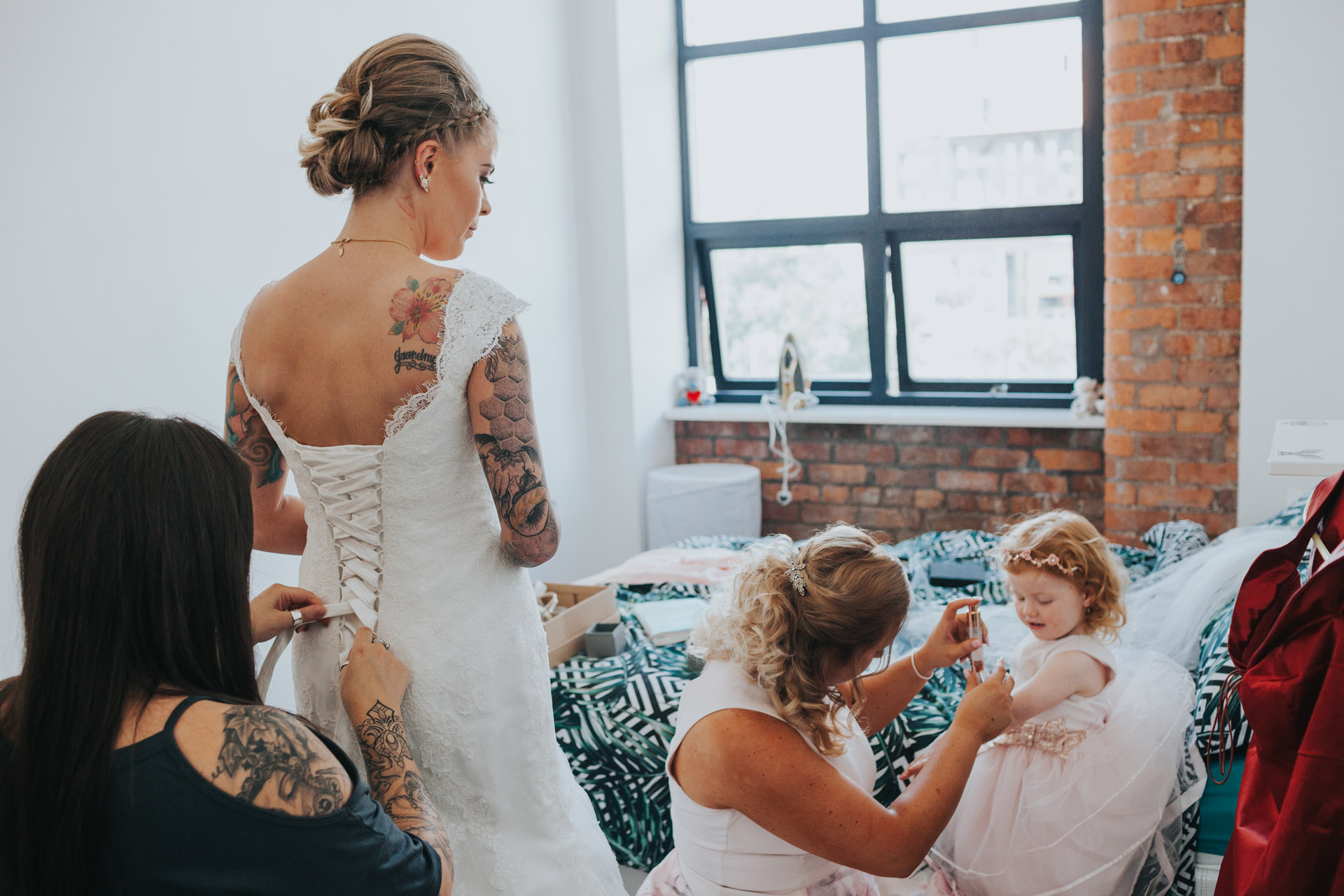 Bride has the back of her wedding dress fastened as bridesmaid puts make up on flower girl.