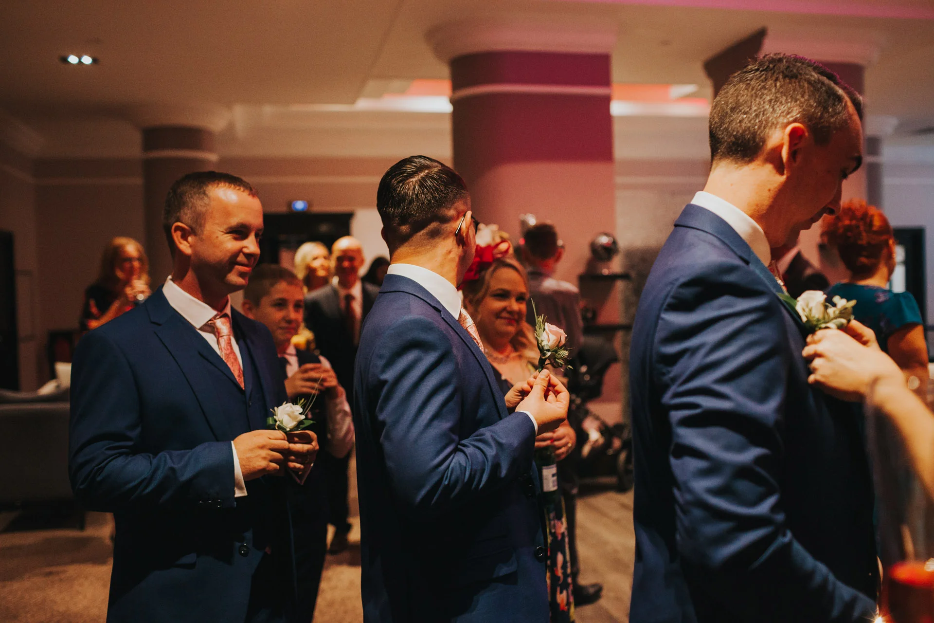 Groom and groomsmen wearing blue suits stand in a line getting their flowers put on.