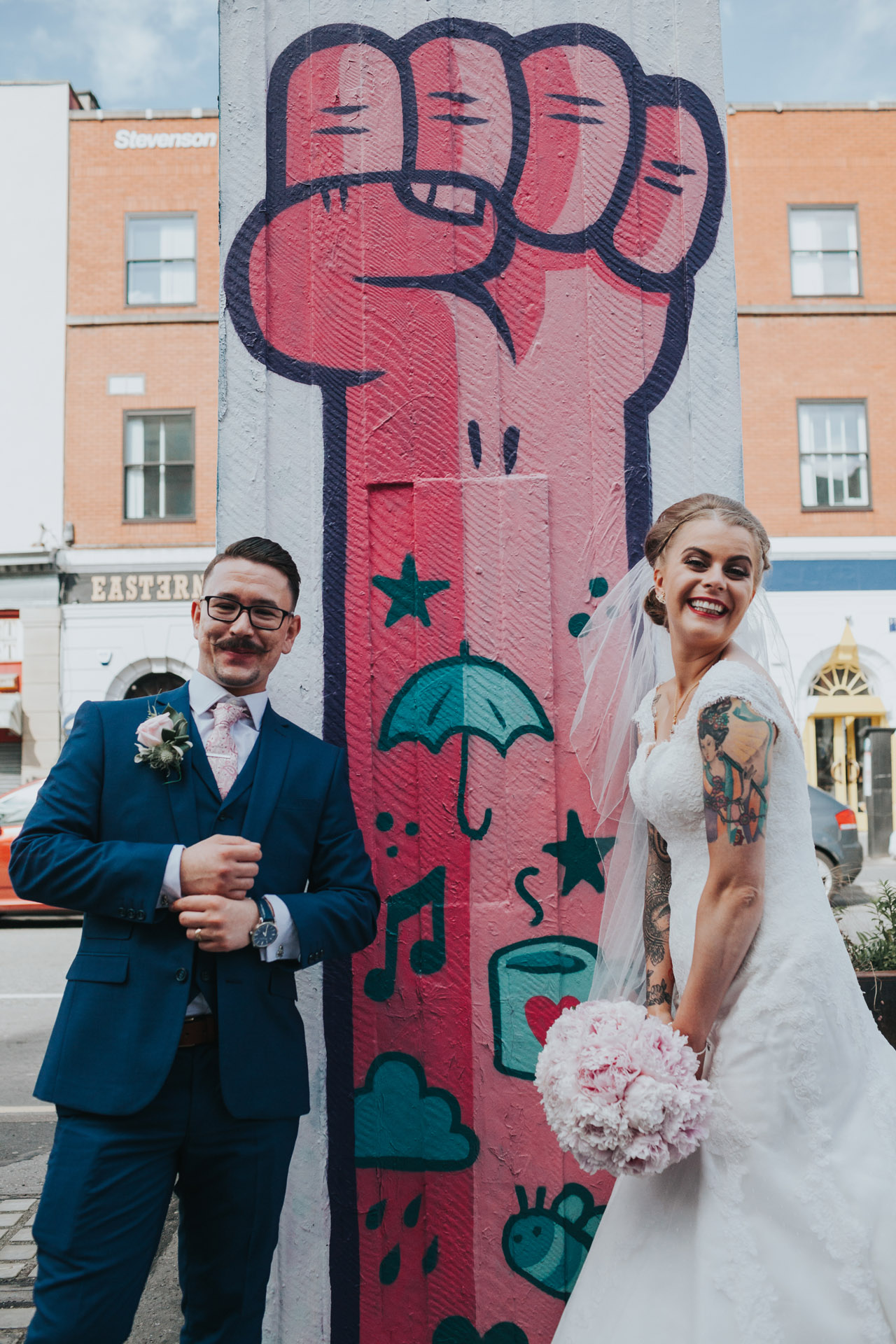 Bride and Groom smile in front of pink power to the people street art in Stevenson Square, Manchester. 