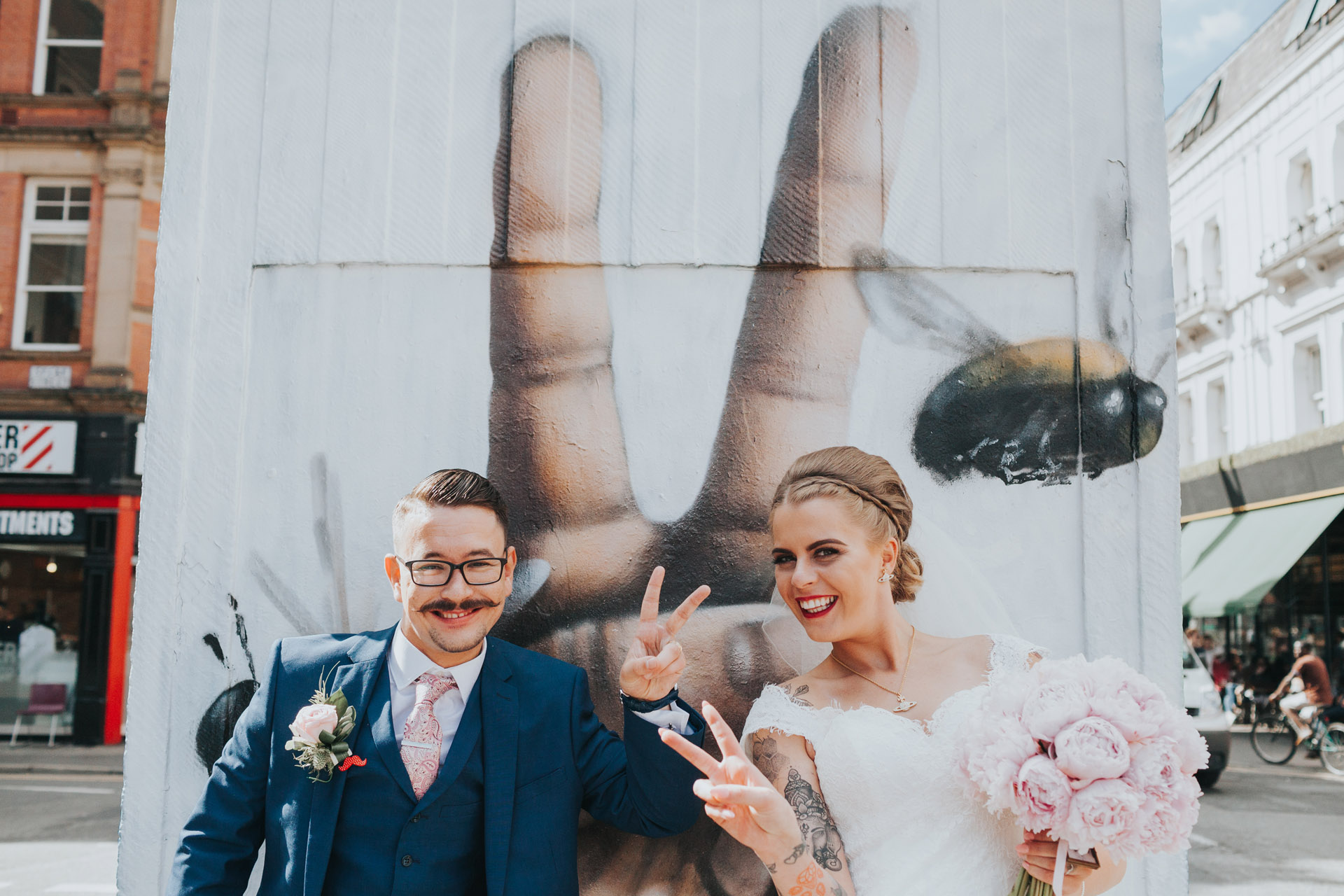 Bride and Groom give the peace sign, in front on Manchester Bee Peace sign Street Art, Stevenson Square, The Northern Quarter, Manchester. 