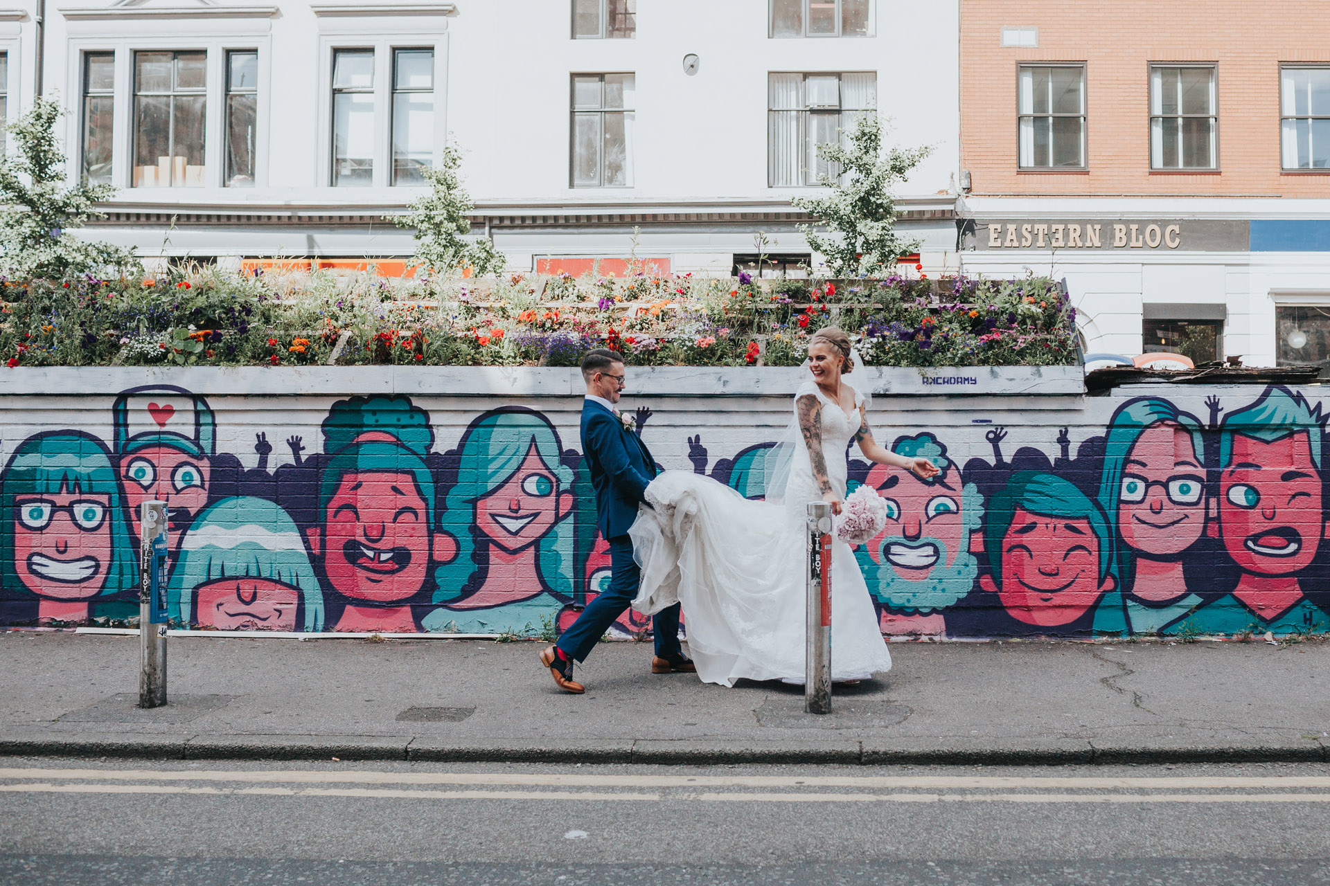 Groom holds brides dress for her as she walks past graffiti, Stevenson Square Manchester. 