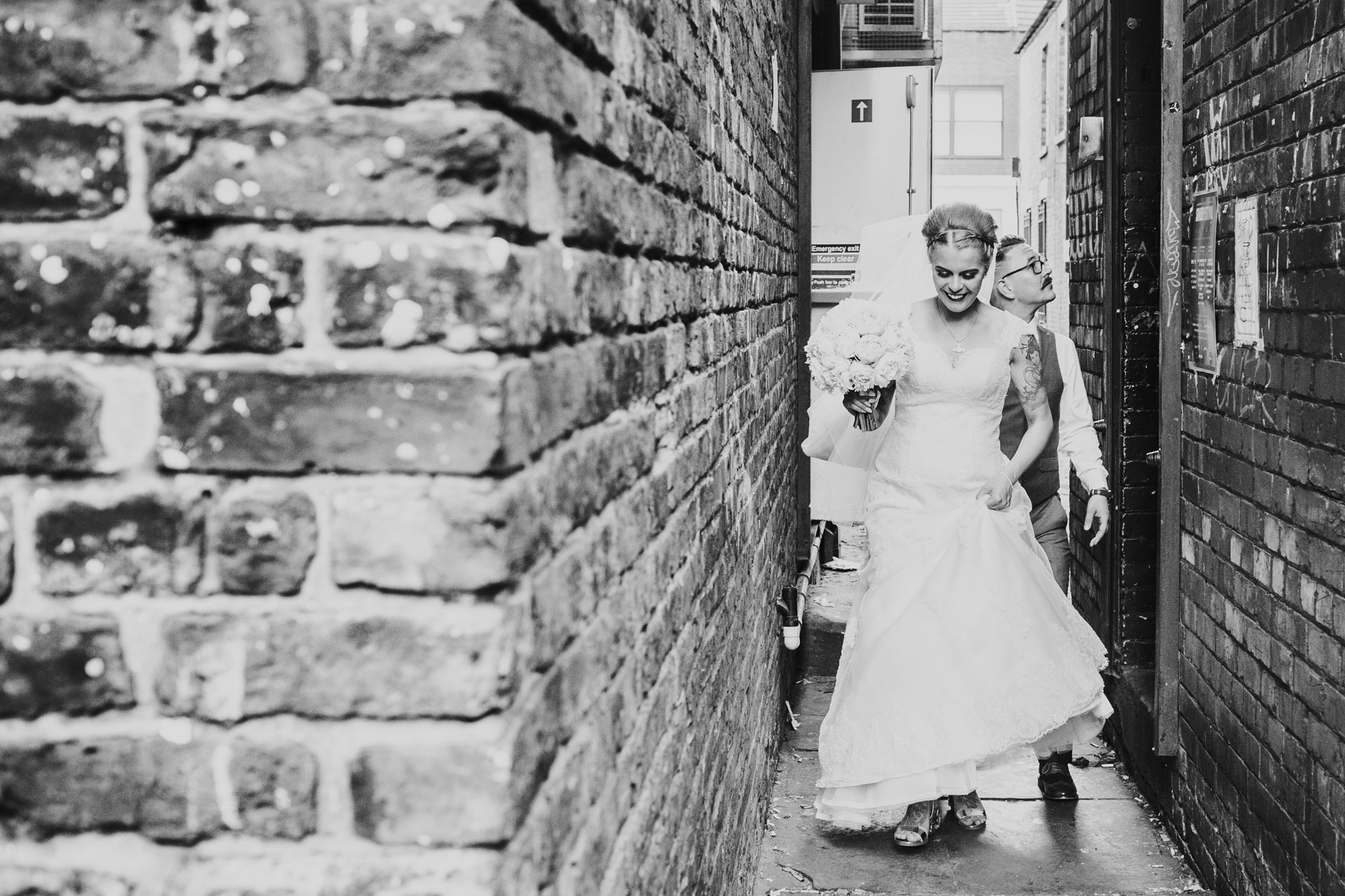 Bride walks down Manchester back alley with her wedding dress and flowers. Photograph in edited black and white. 