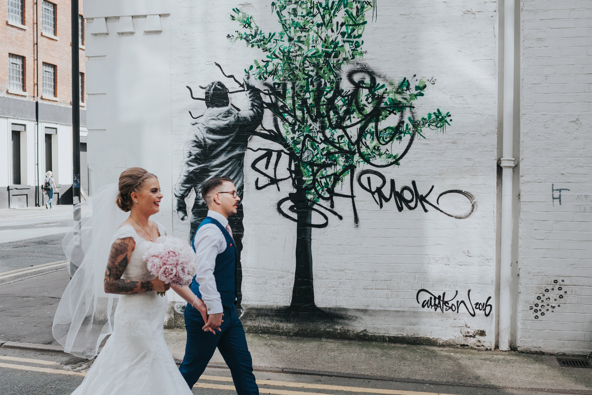 Bride and Groom walk past tree graffiti on Faraday Street, Manchester. Photograph processed in colour. 