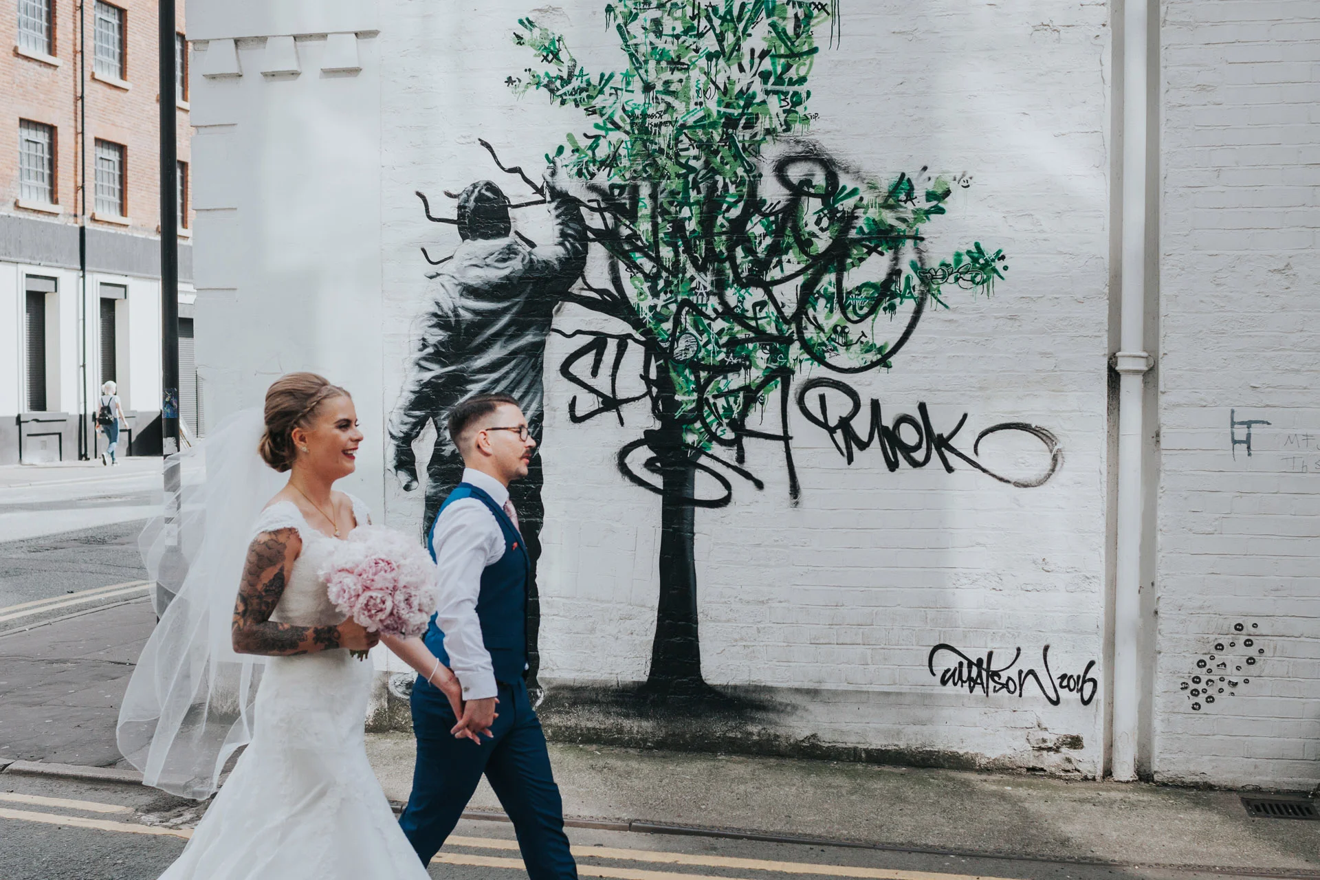Bride and Groom walk past tree graffiti on Faraday Street, Manchester. Photograph processed in colour.