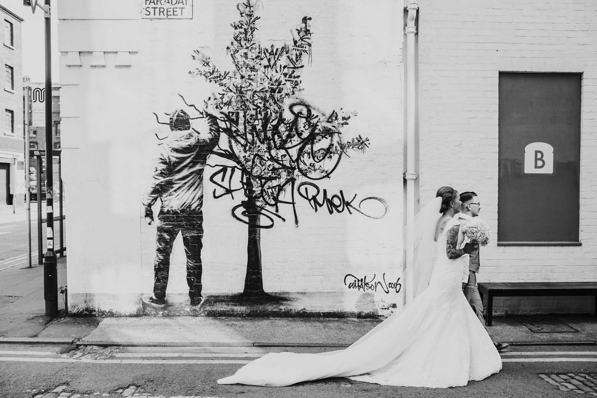 Photograph processed in black and white. Couple walk by Faraday Street, Tree Graffiti in Manchester. 