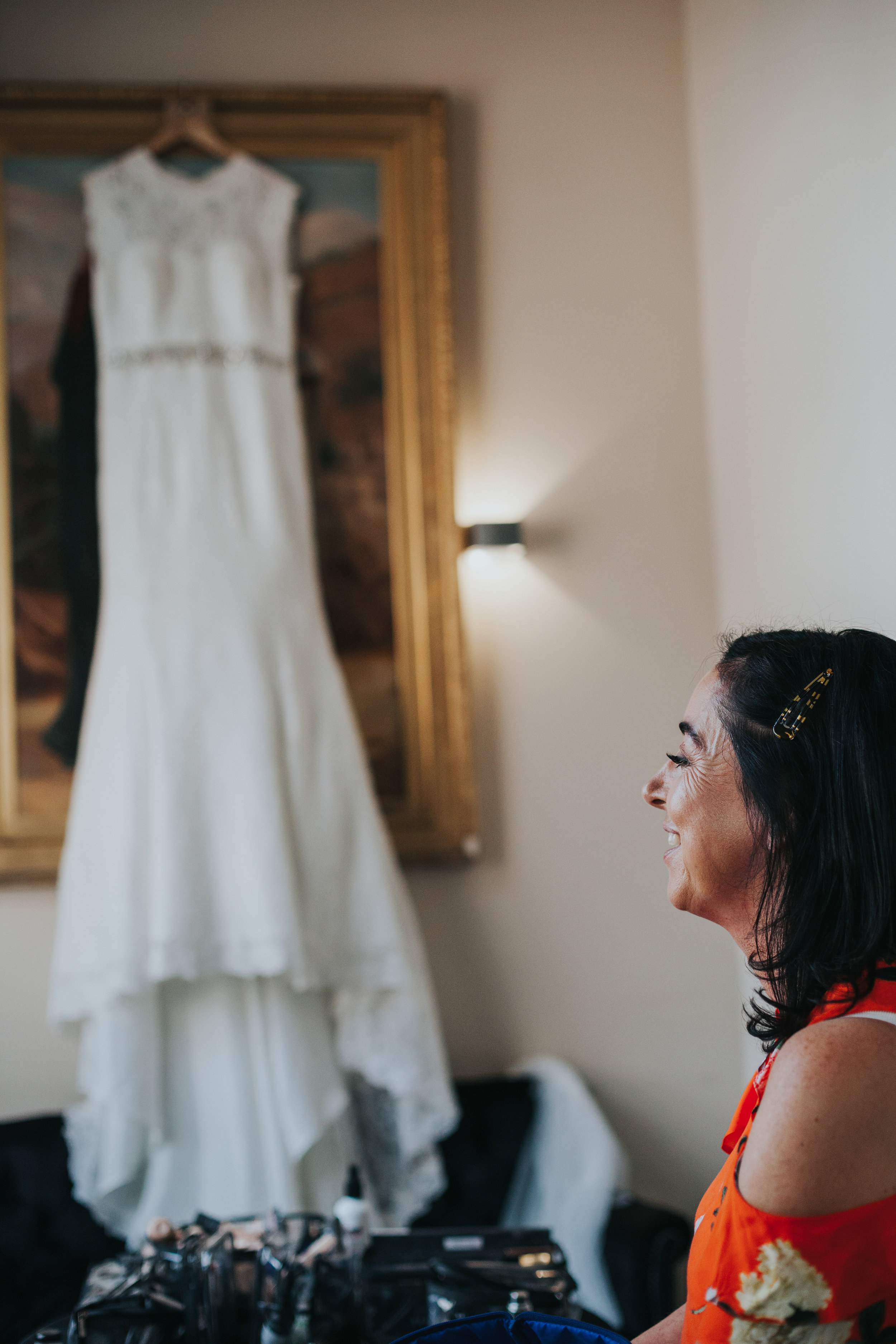 Brides mother stands in front of her wedding dress smiling. 