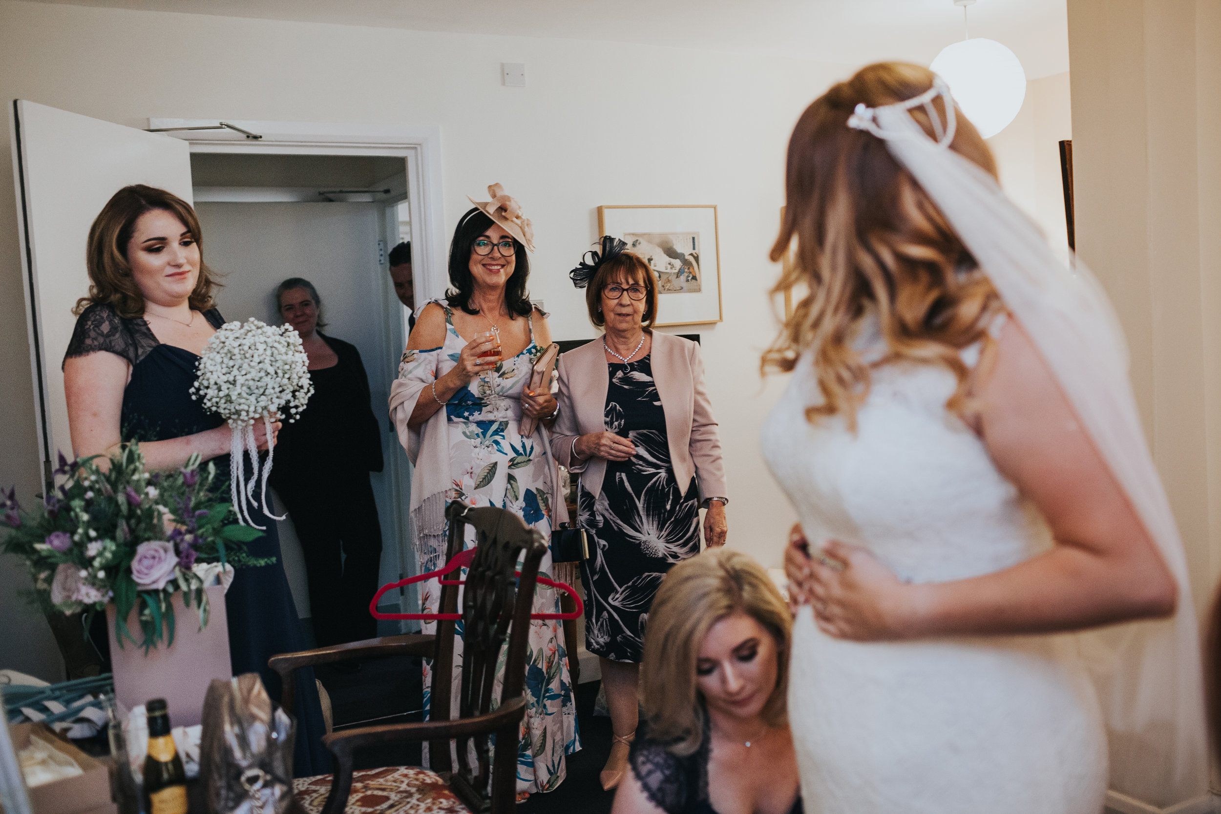 Brides family and bridesmaids smile as they look at the bride in her dress. 