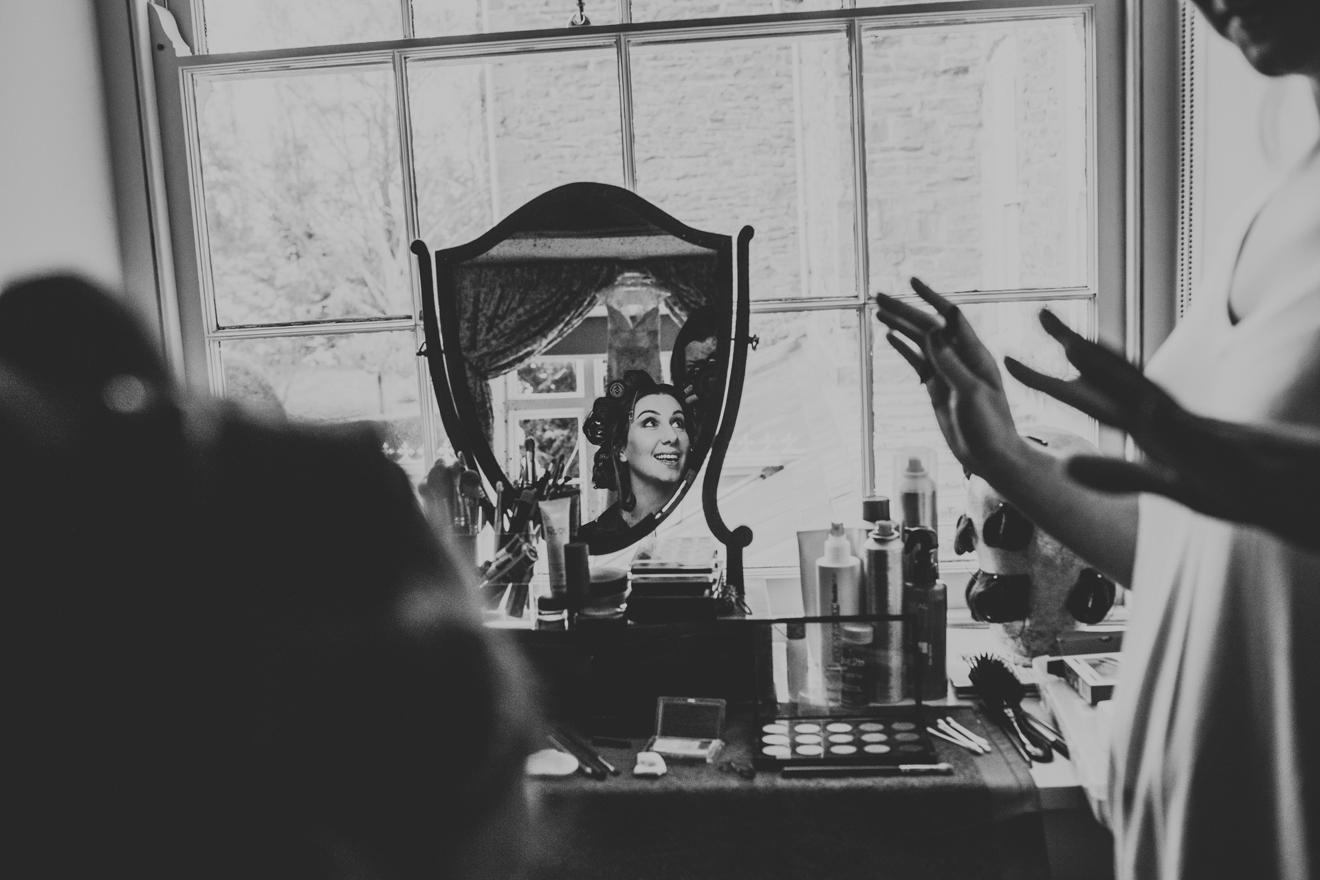 The brides reflection in the mirror as she smiles at her friend, photograph in black and white.