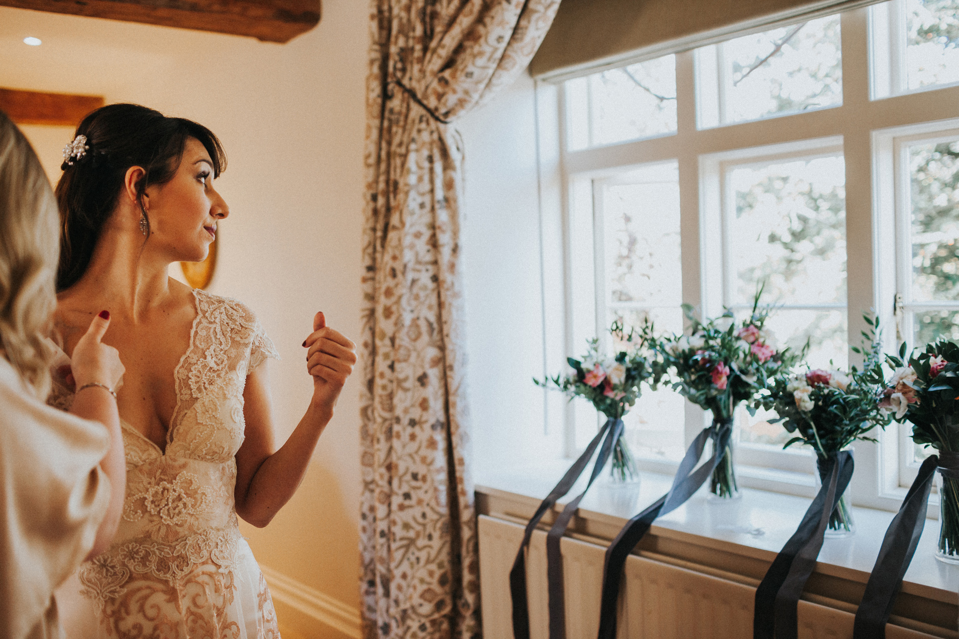 Bride looks out of the window over the wedding flowers as her bridesmaids help her with her dress. 