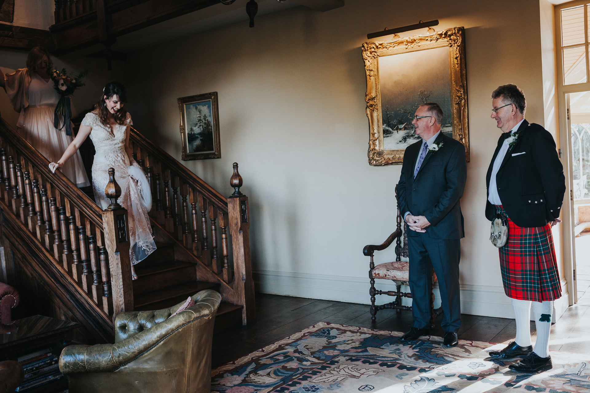 The brides fathers watch her coming down the stairs in her wedding dress. 