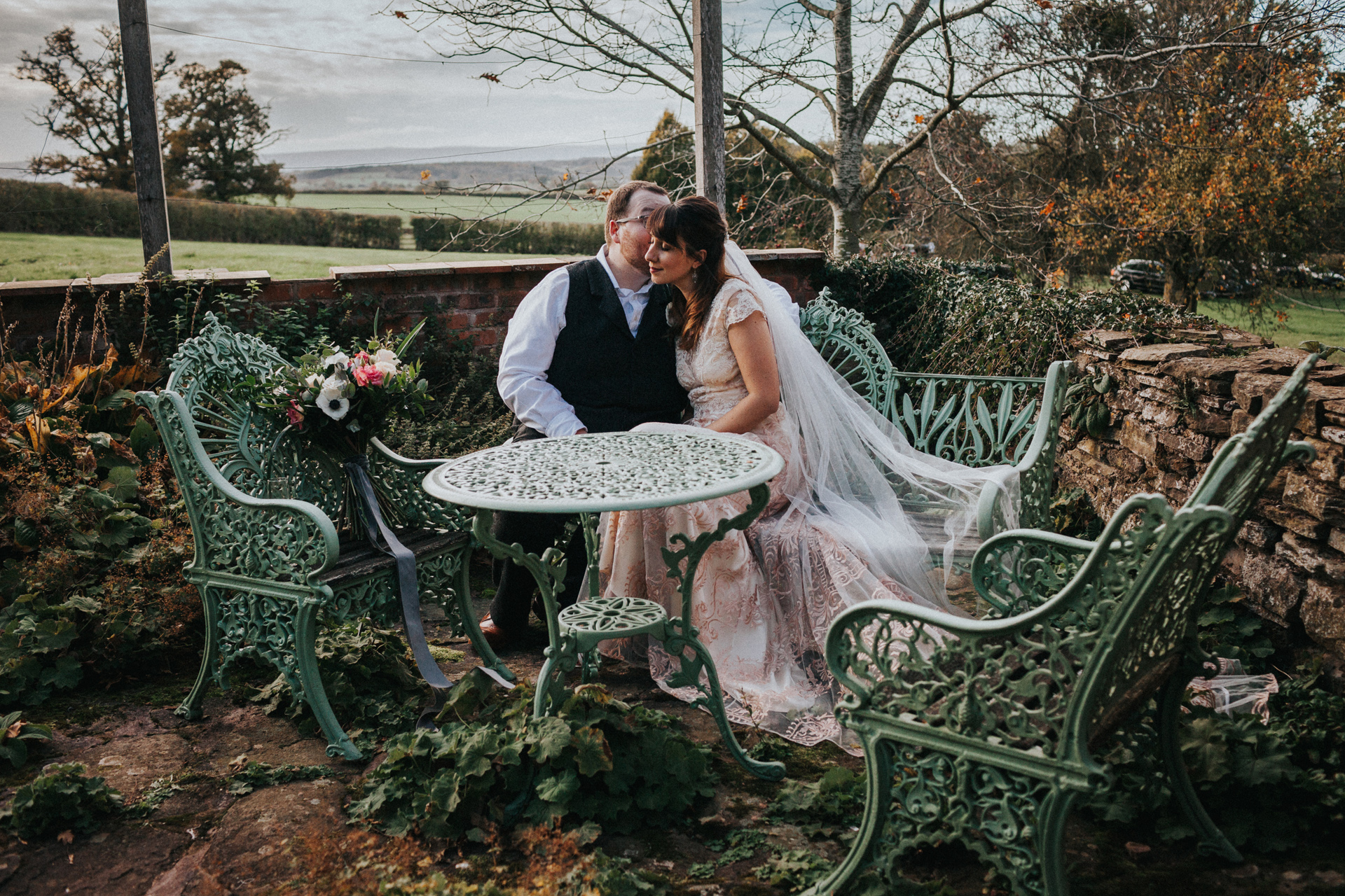Bride and groom share a moment on teal iron chairs at Dewsall Court.