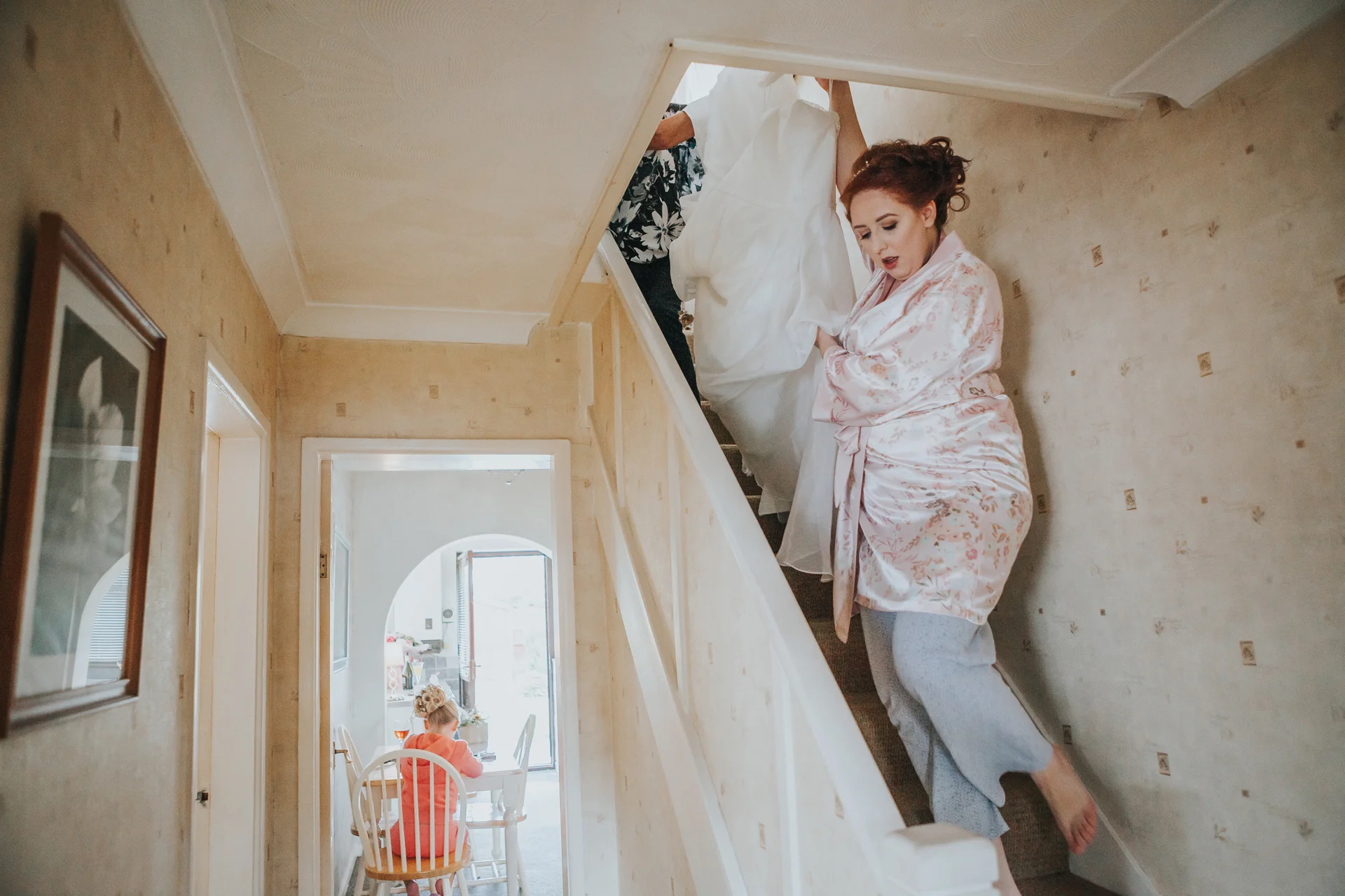 Bride bringing her dress down the stairs, bridesmaid waits in kitchen.