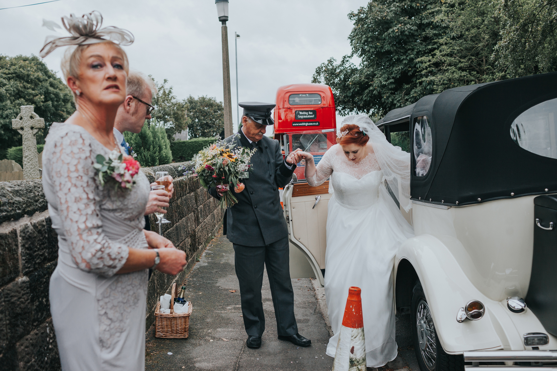Brides mother waits outside wedding car as bride is helped into it by driver, big red bus in the back ground. 