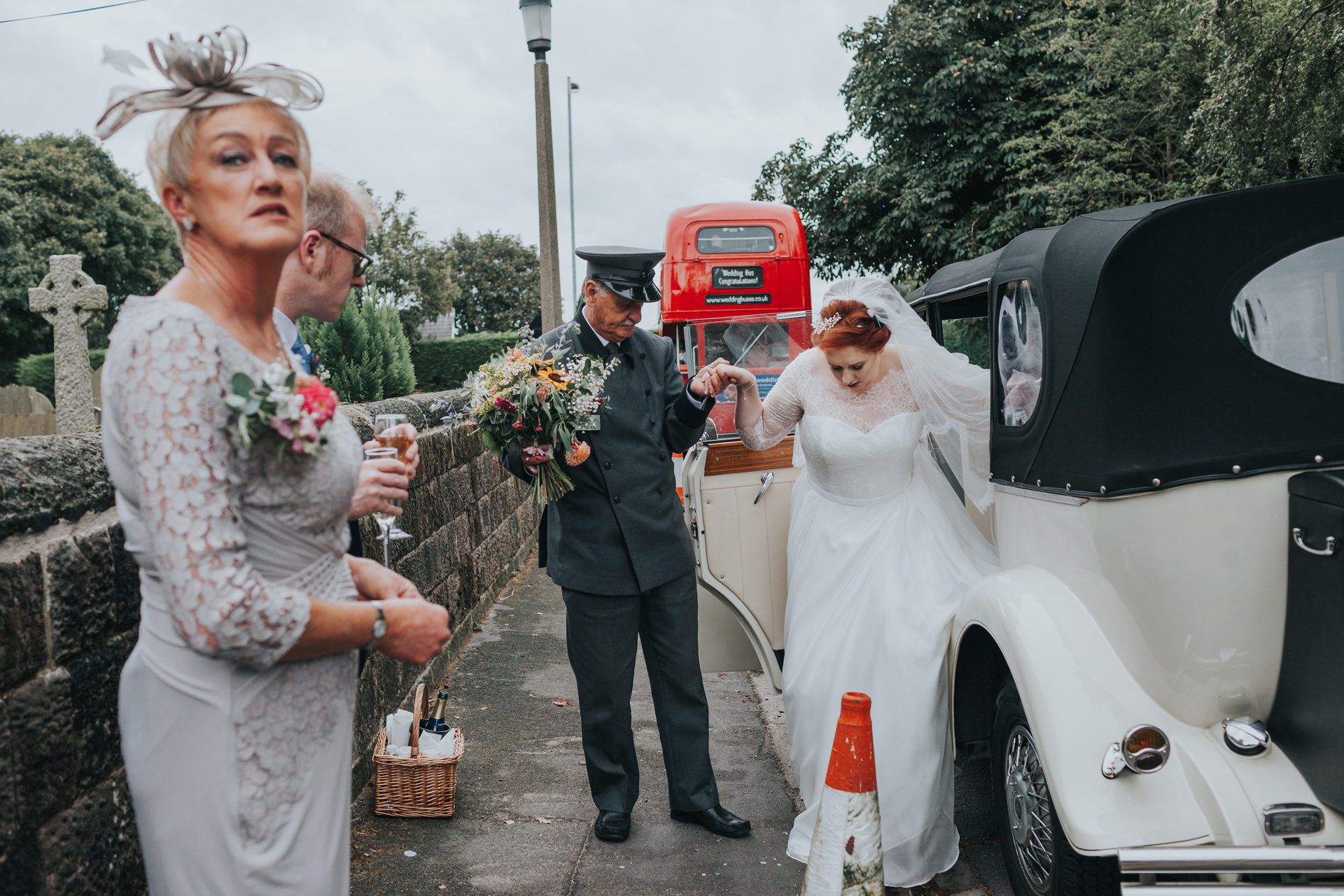 Brides mother waits outside wedding car as bride is helped into it by driver, big red bus in the back ground.