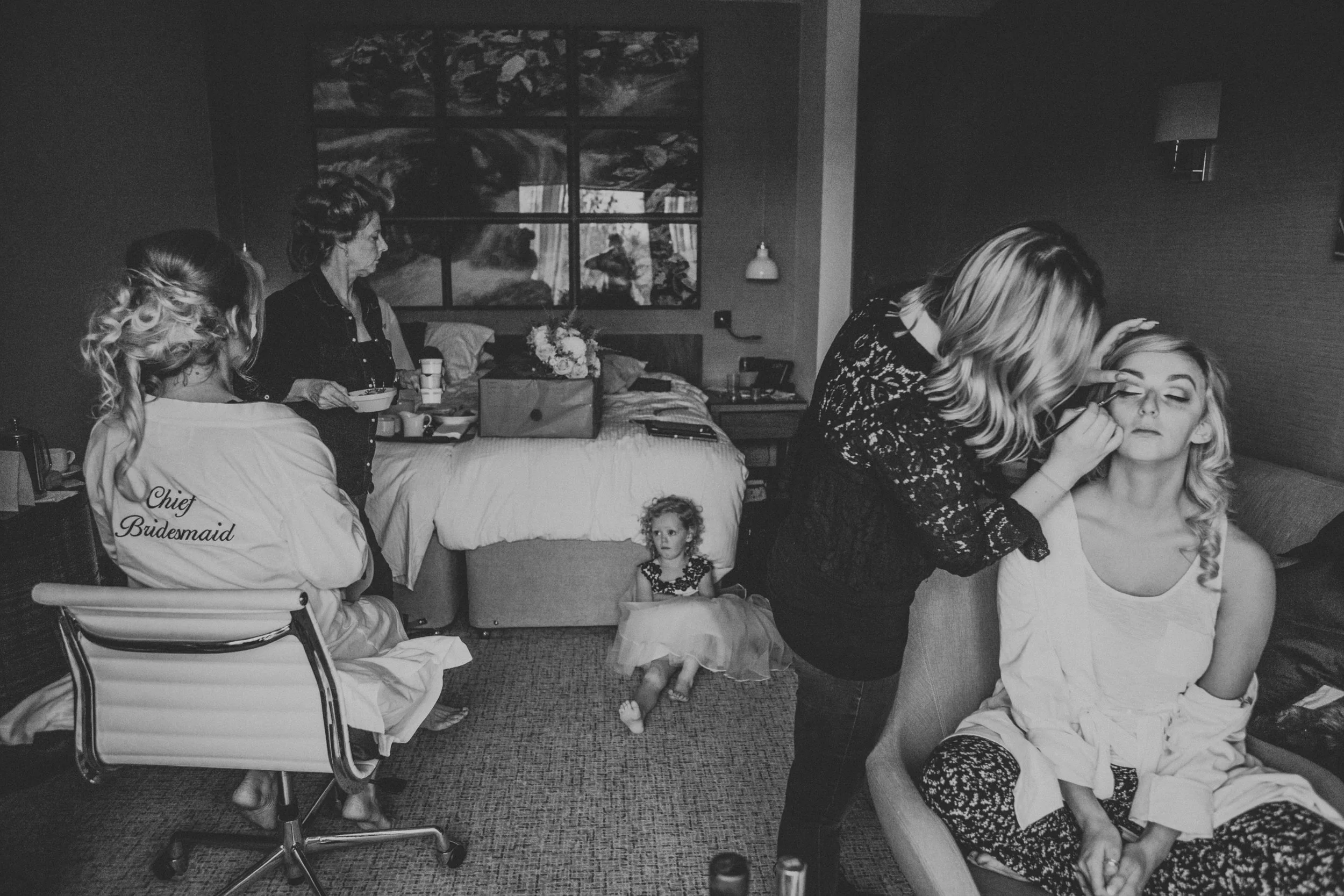 Bride getting her make up done, as others get ready and flower girl sits on the floor. 