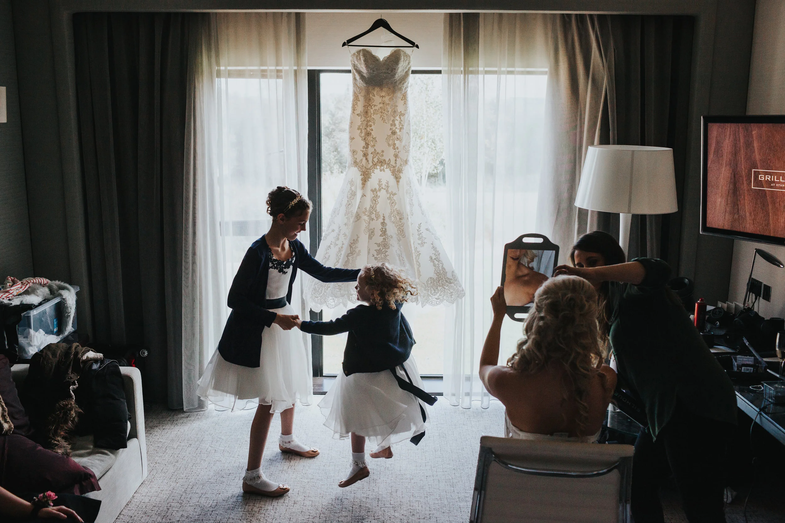 Flower girls dance in front of wedding dress as bride has the finishing touches to her make up done. 