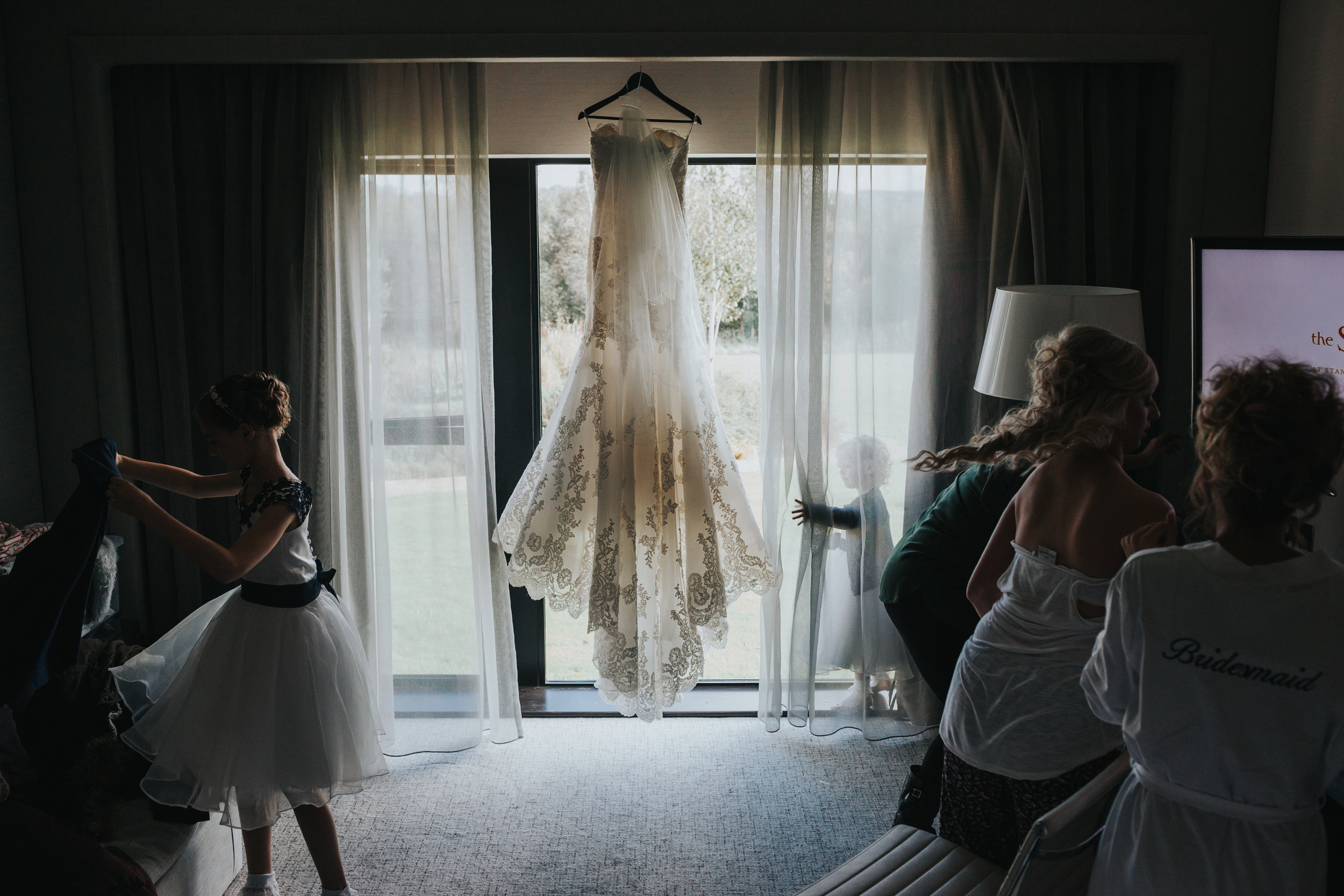 Dress hangs in back ground as bridal party continue to get ready in the shadows. 