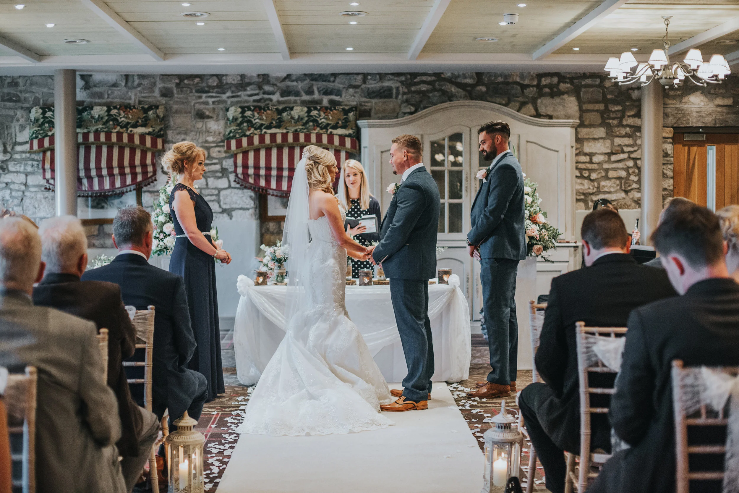 Shot from the back of the room. Bride and groom hold hands. 