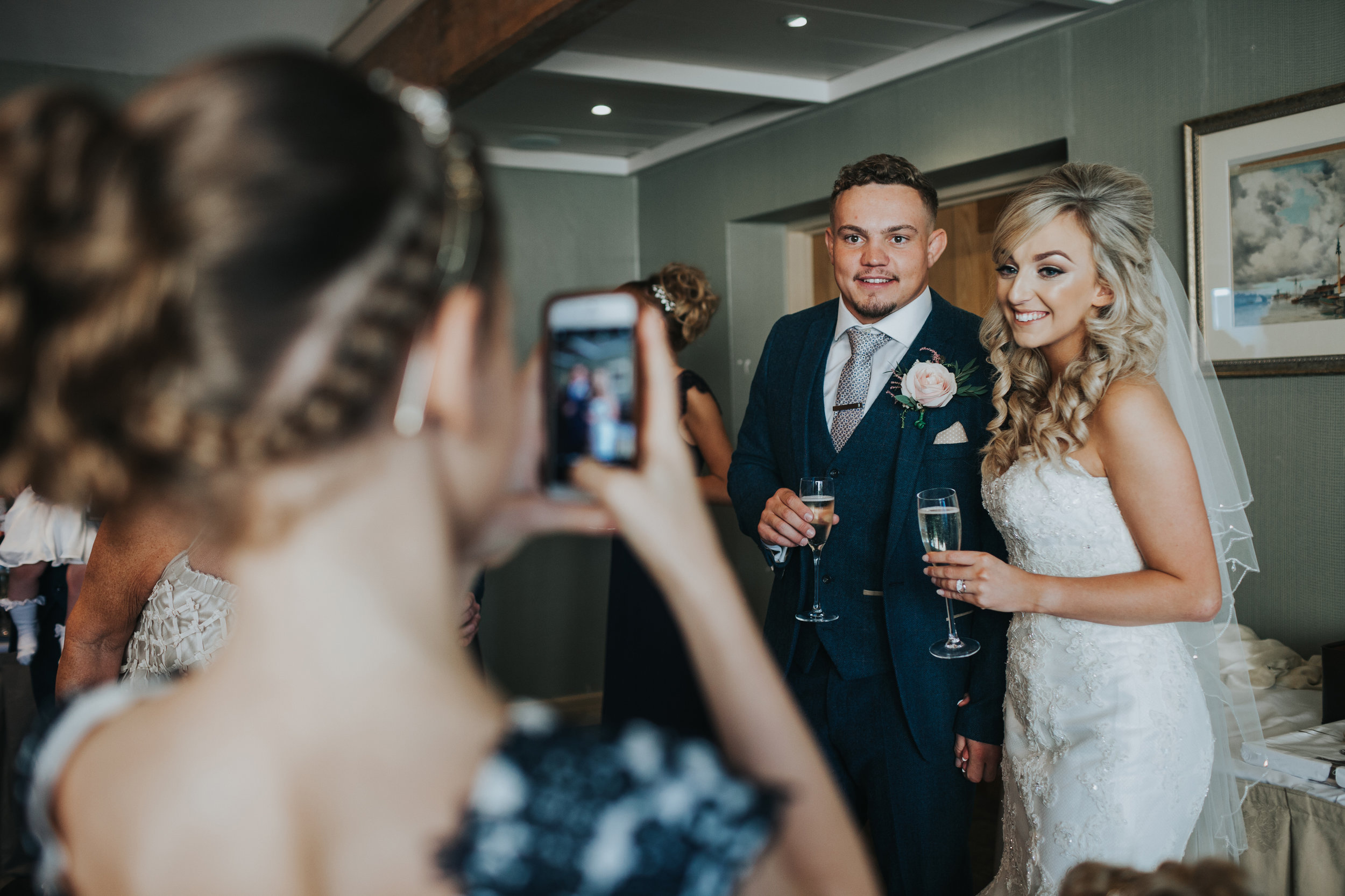 Bride and groom pose for their flower girl to take a photograph on her phone.