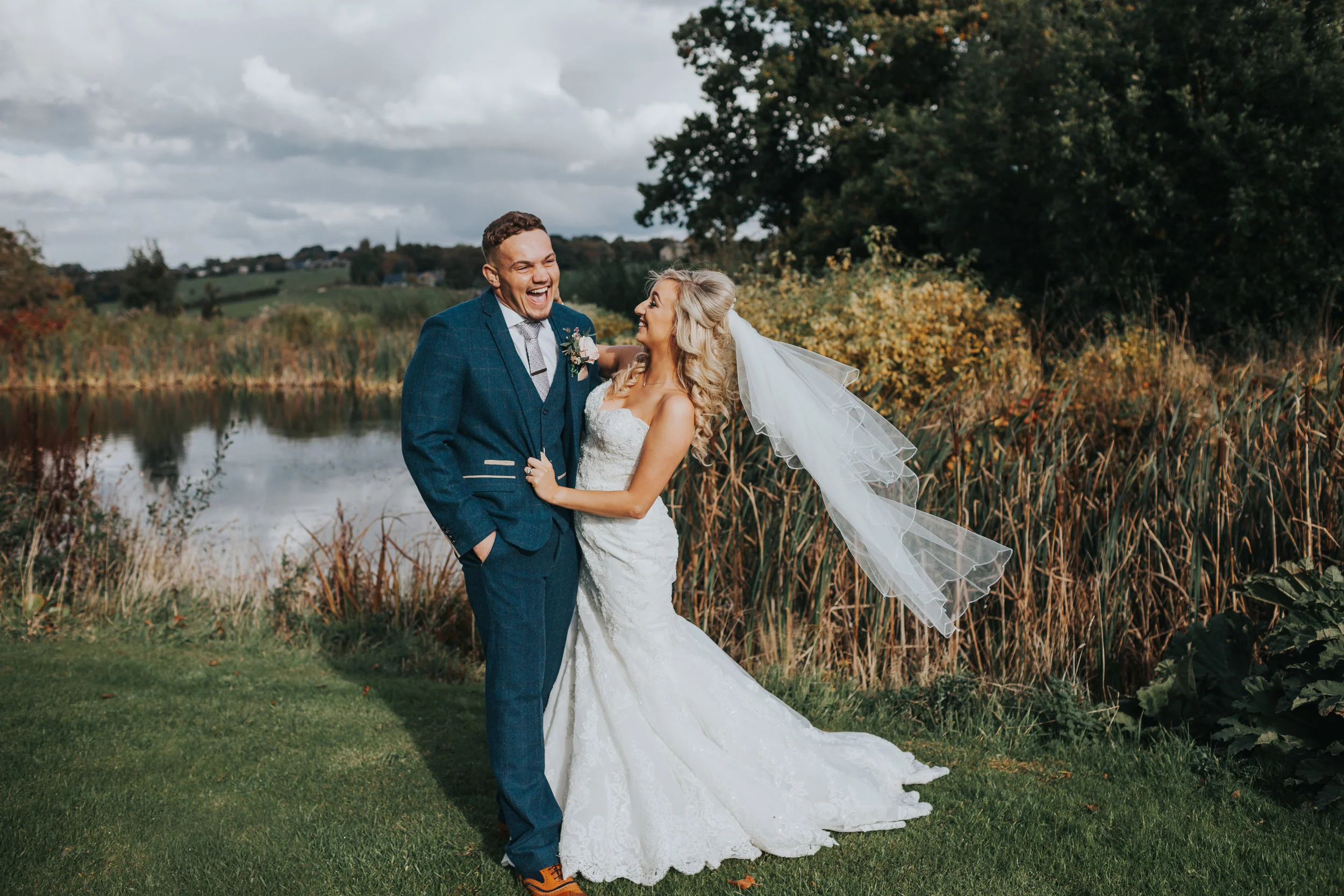 Bride and groom laugh together in front of pond.