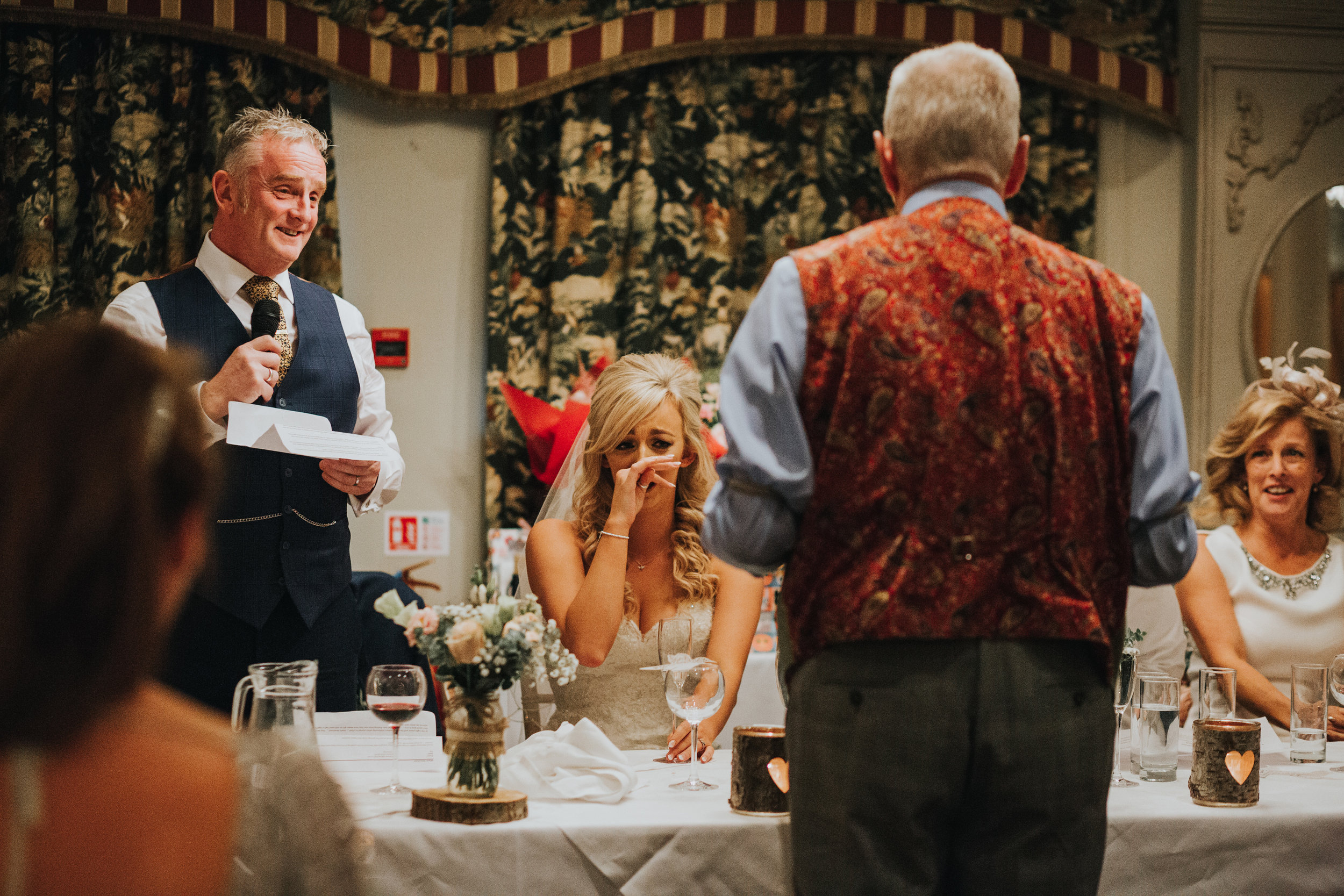 Brides uncle approaches table and bride holds hand over her mouth laughing and looking scared. 