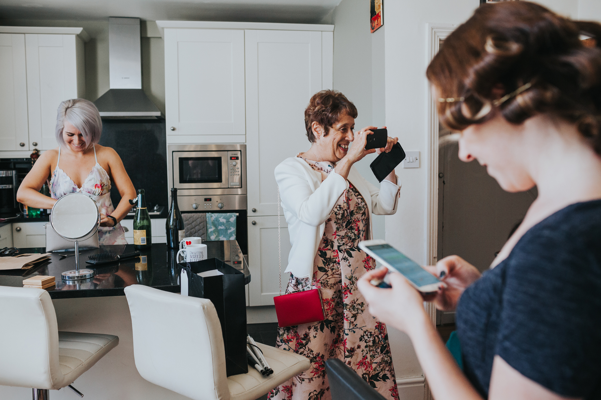 Bride, her mum and a bridesmaid hand out in the kitchen together taking photographs with their phones. 