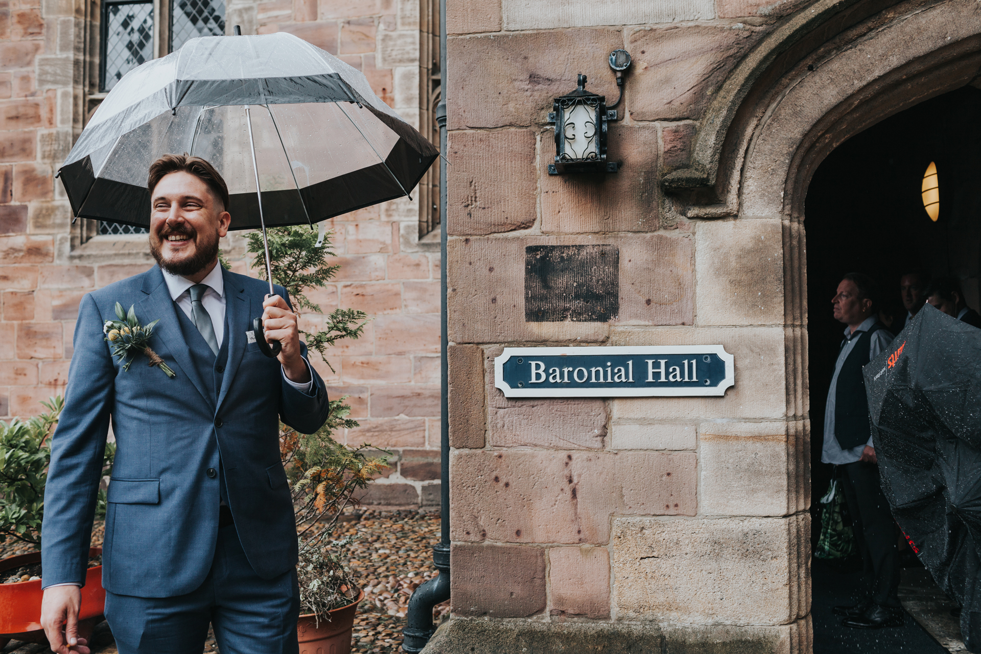 Groom welcomes wedding guests under umbrella outside Chetham Library.