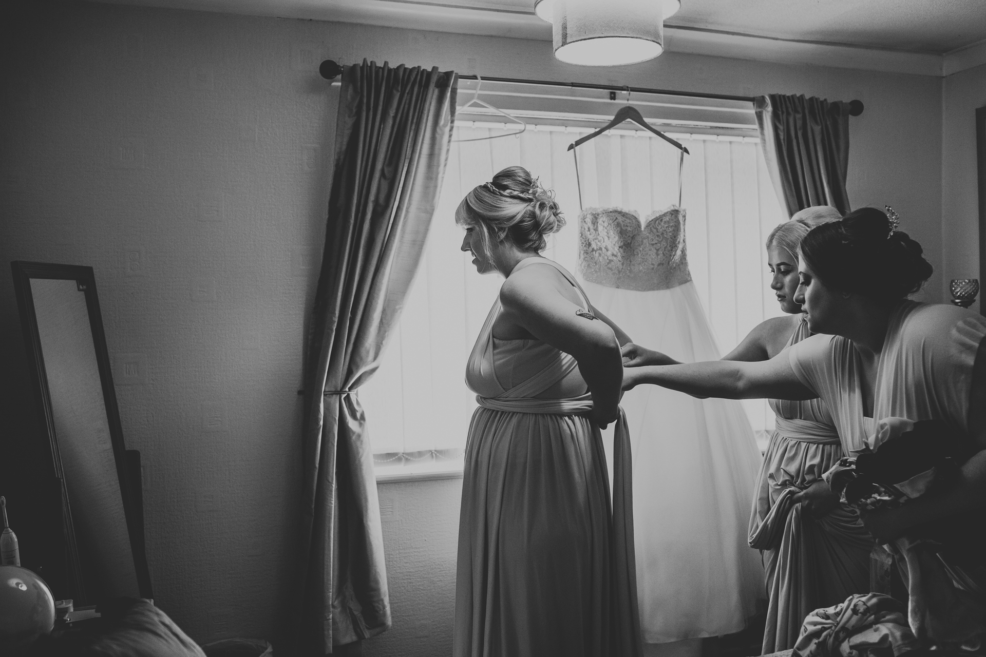 Bridesmaids help to dress another in front of a window, the wedding dress hangs from the curtain rail above the window.