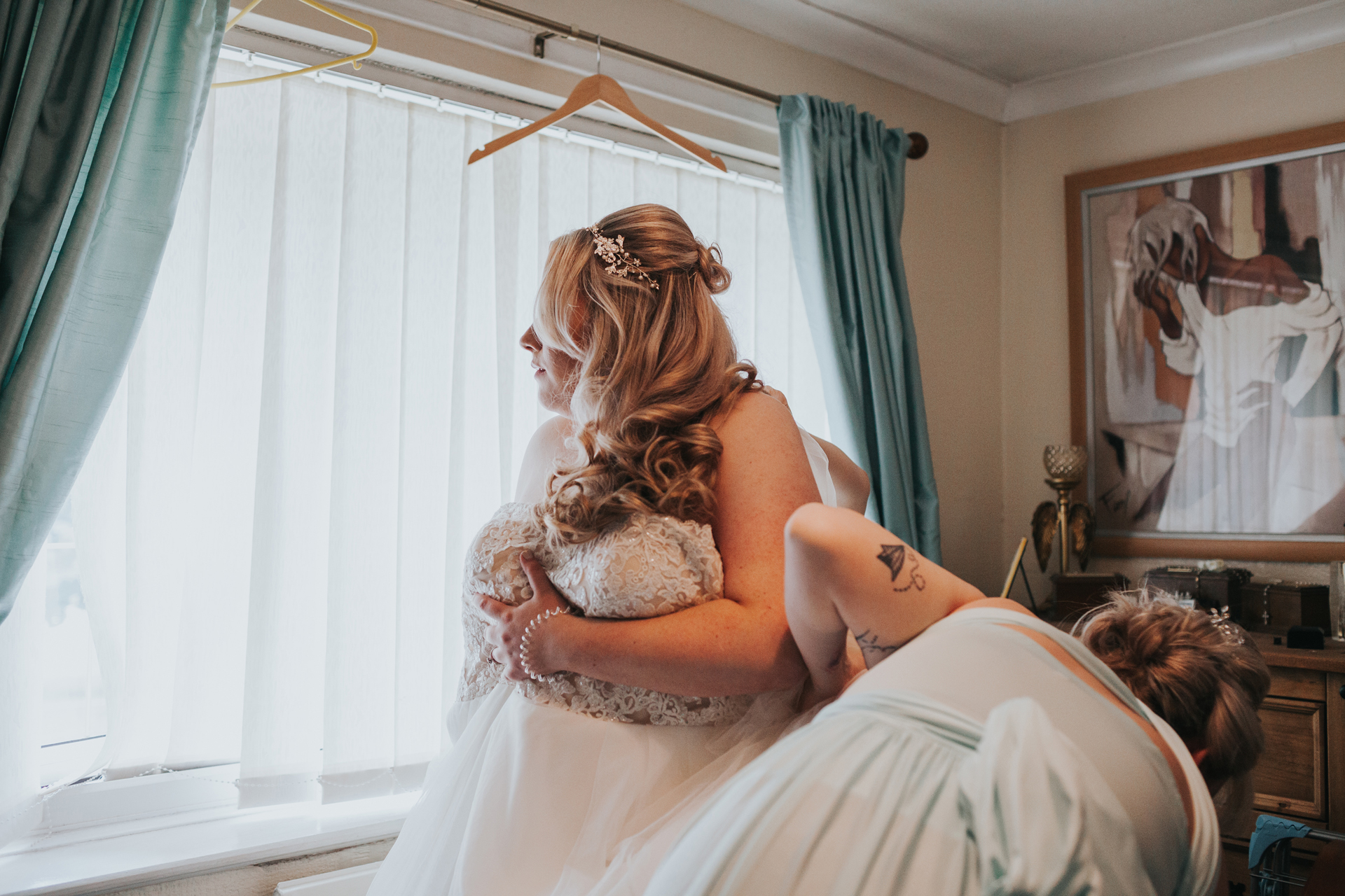 Bride looks out of the window and her bridesmaids help fix her dress. 