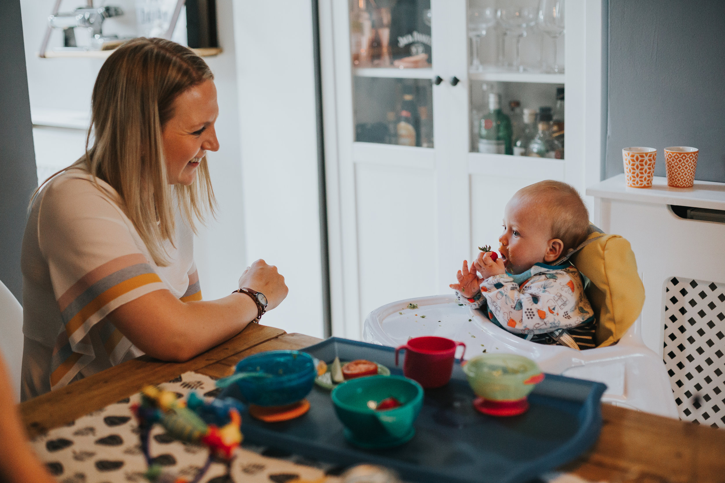 Mum watches baby eating smiling. 