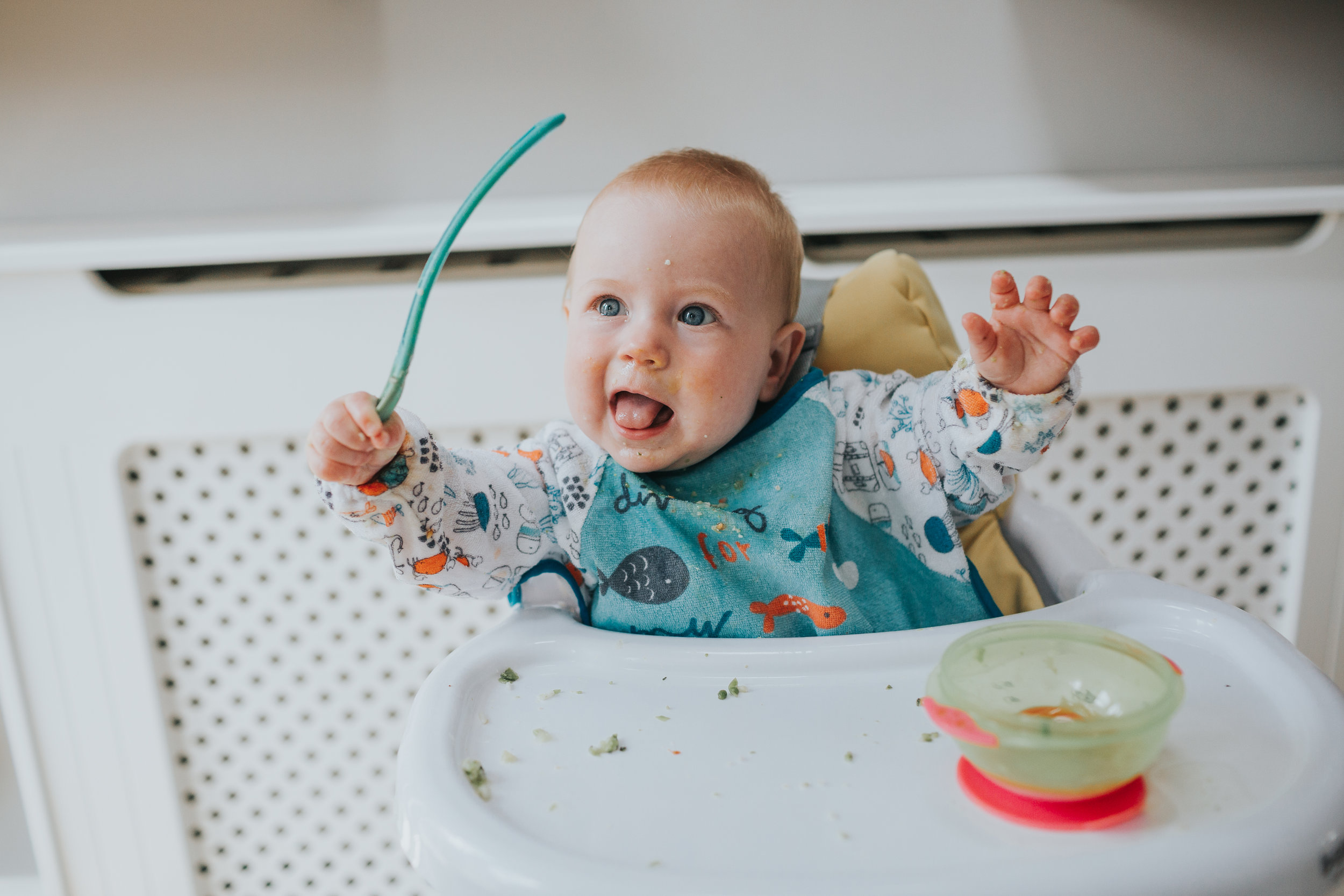 Baby pulls tongue out and waves his spoon in the air.