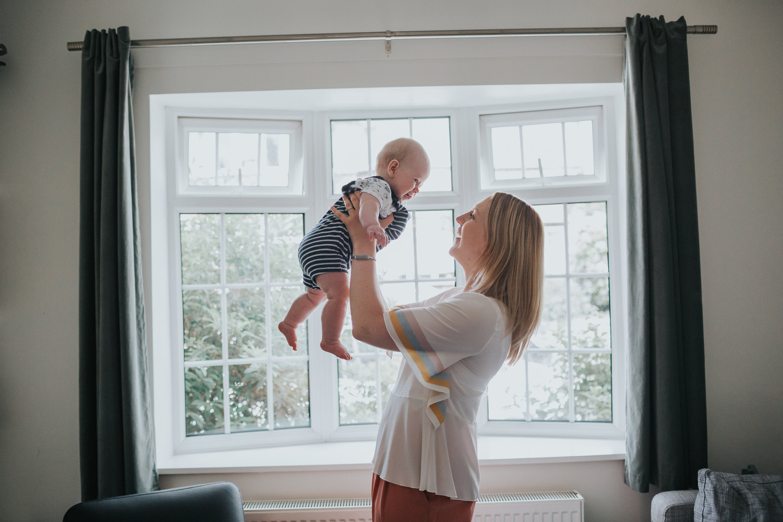 Mum chucks baby up and down in front of their living room window. 