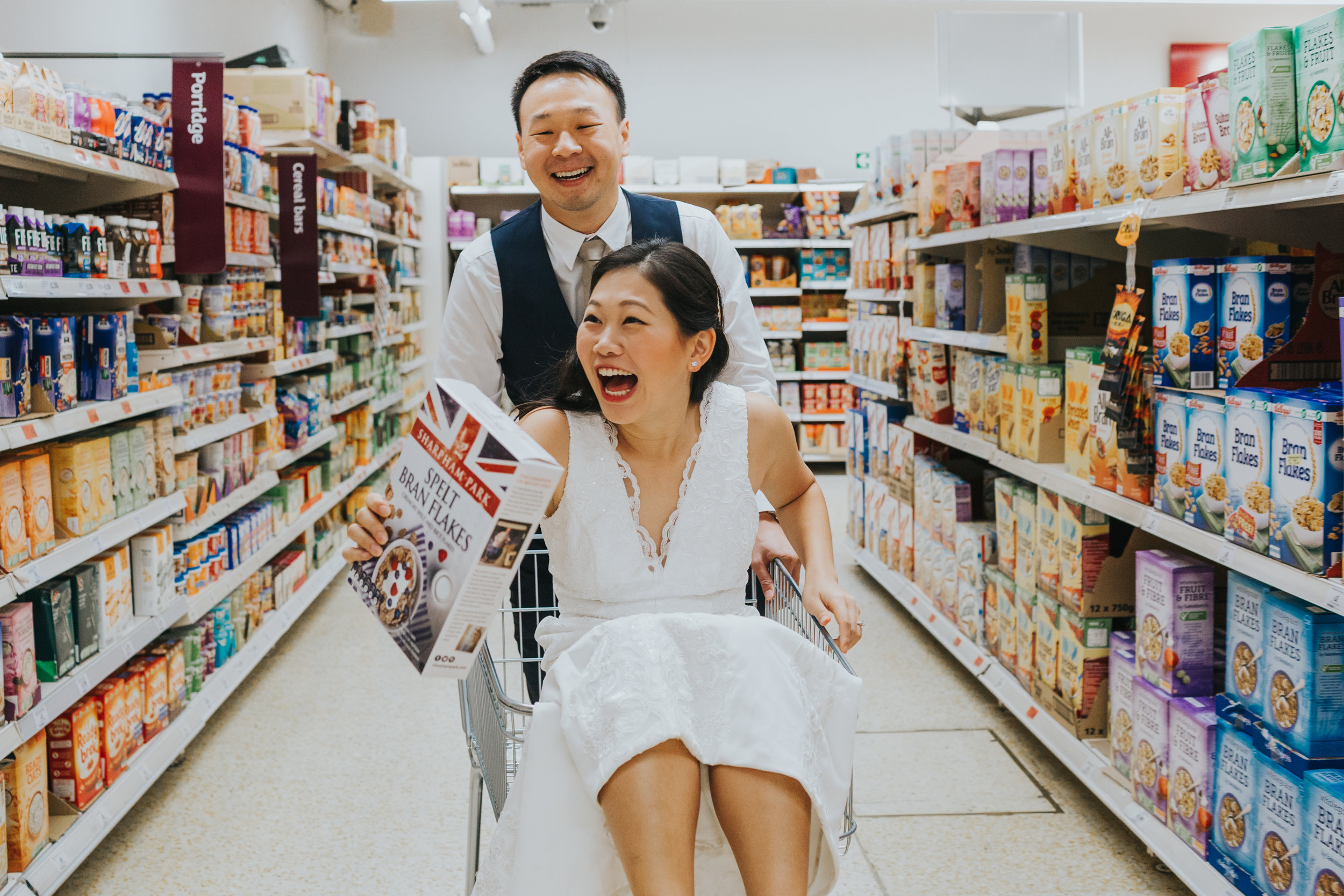 Groom pushes bride around in shopping trolly. 