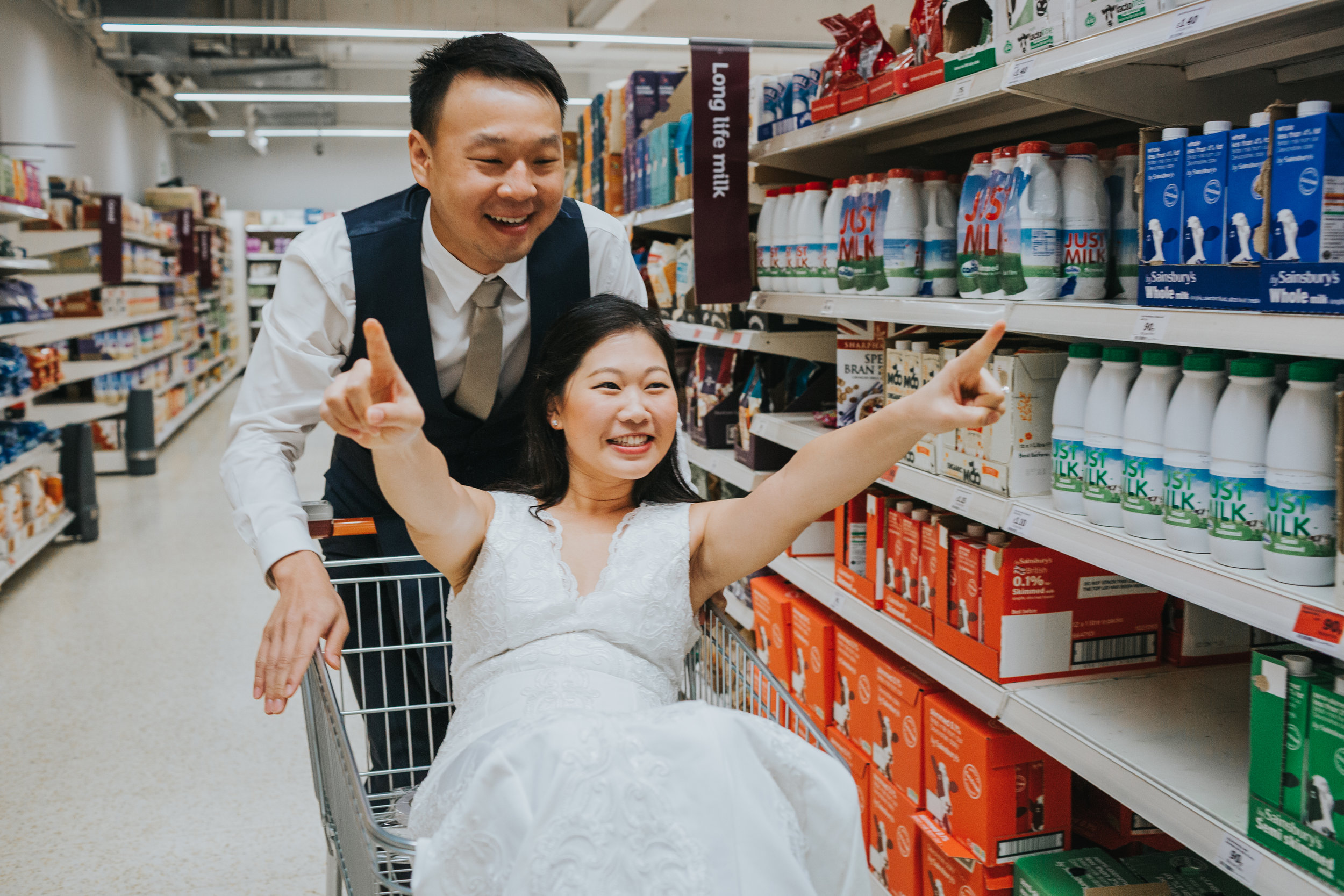 Bride poses in rock and roll pose as Groom pushes her down the aisle in shopping trolley. 