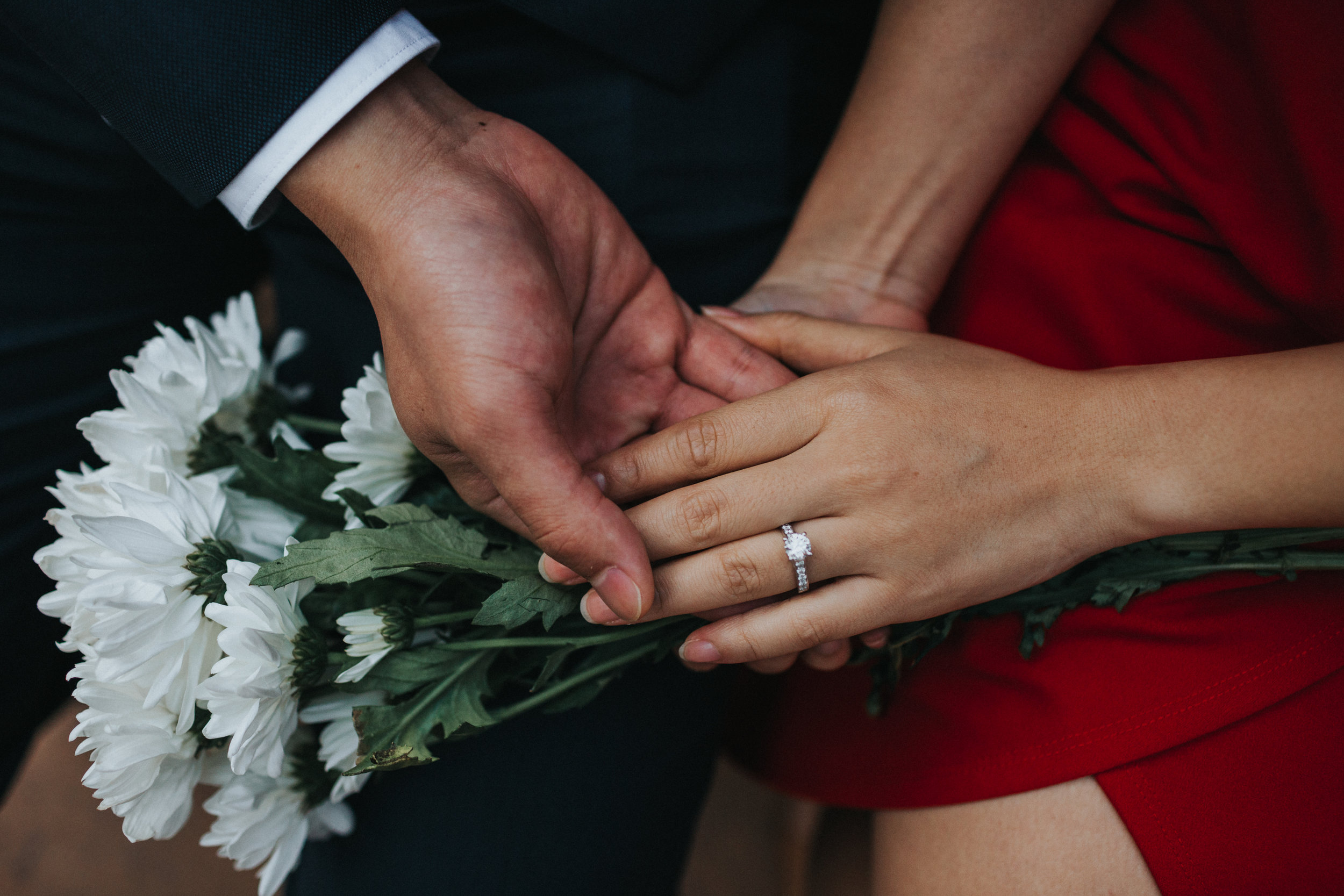 Couple hold hands, close up of engagement ring on Cristal's Red dress and white flowers in the back ground. 