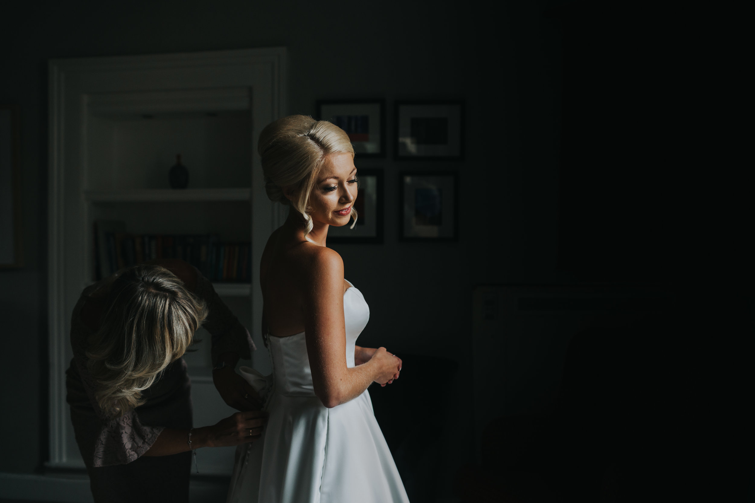 The bride stands in window light as her mother helps her put on her dress. (Copy)