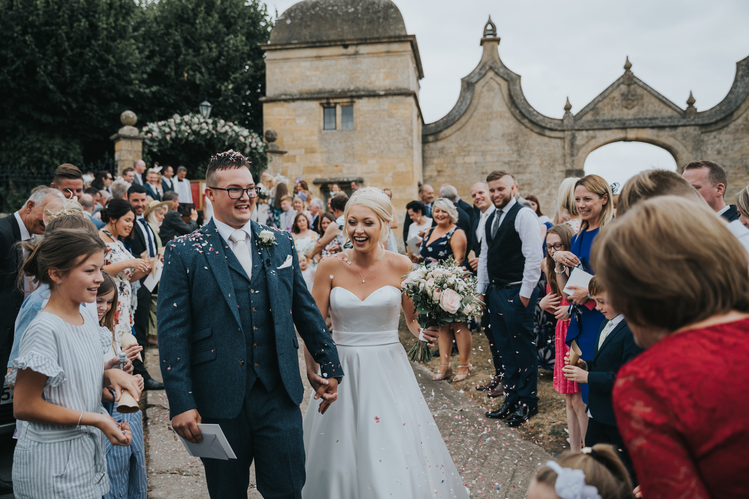 Bride and Groom smiling surrounded by guests and covered in confetti.