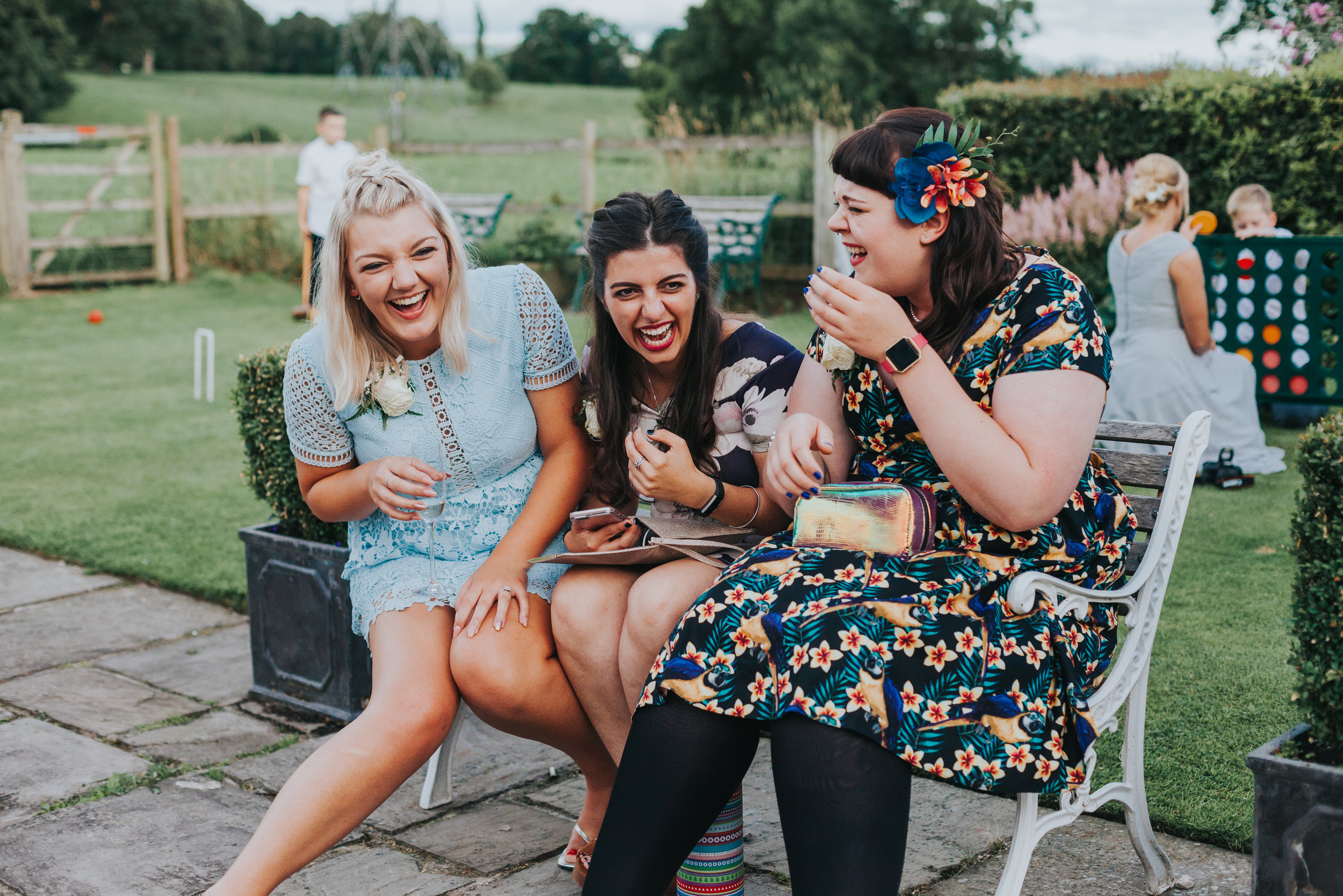 Candid shot of 3 female wedding guests dressed in brightly coloured clothes sit on a bench and laugh together at a wedding in Manchester, while children play lawn games behind them. 