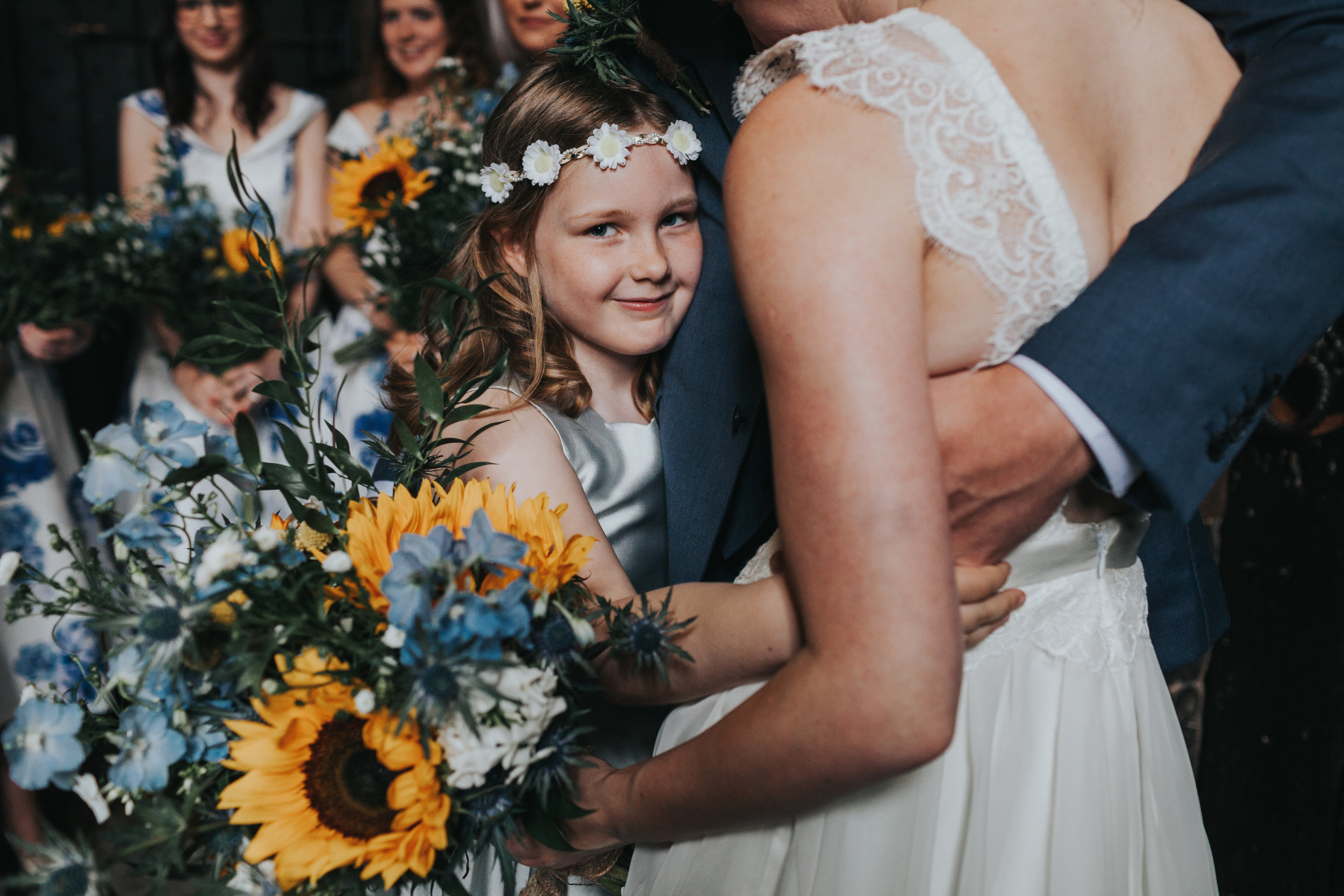 Unposed photograph of a flower girl hugs the bride and groom while looking into the camera at Chetham Library Manchester.