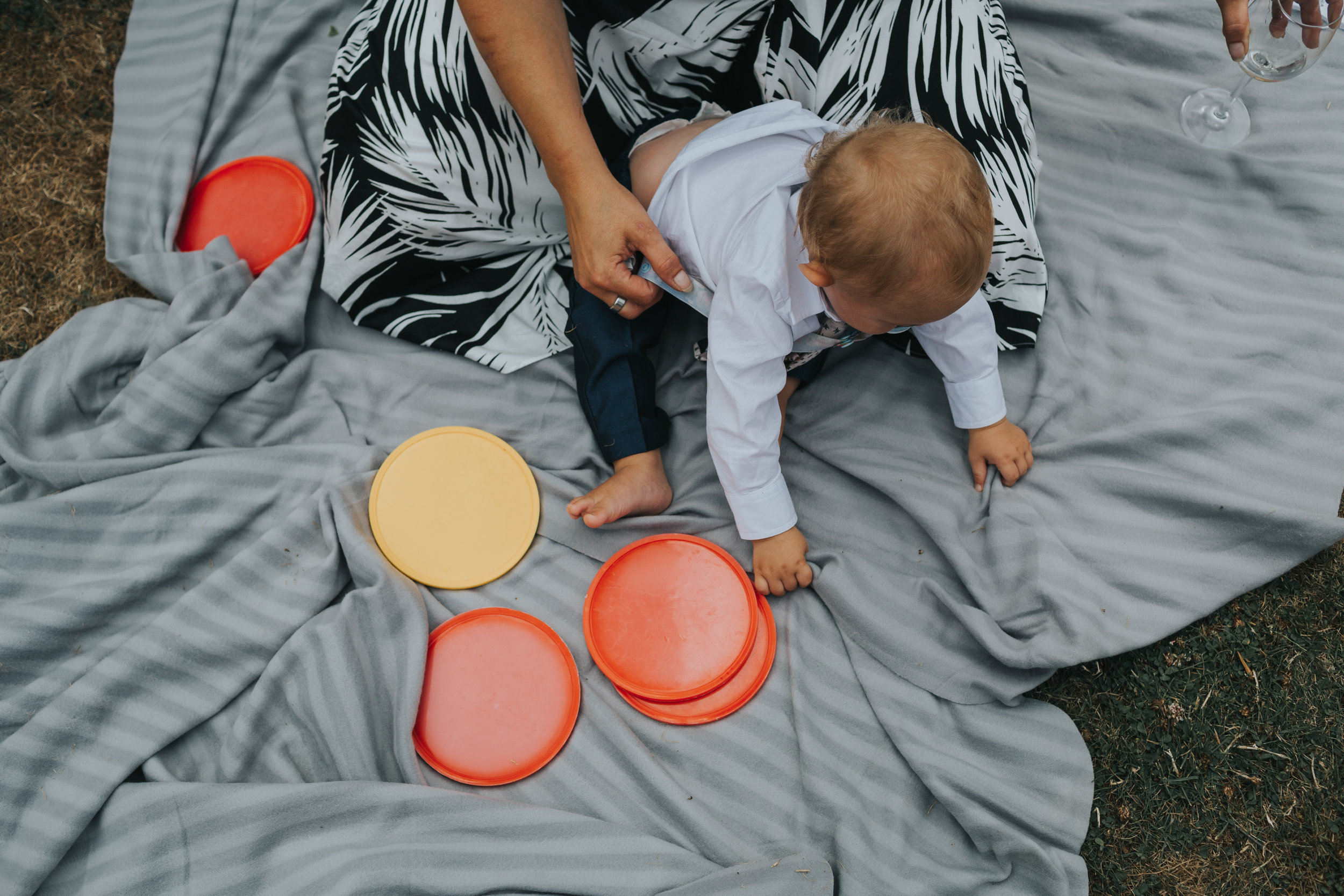 6 month old child photographed from above plays with connect 4 pieces on a grey blanket at an outdoor wedding in Manchester, UK. 
