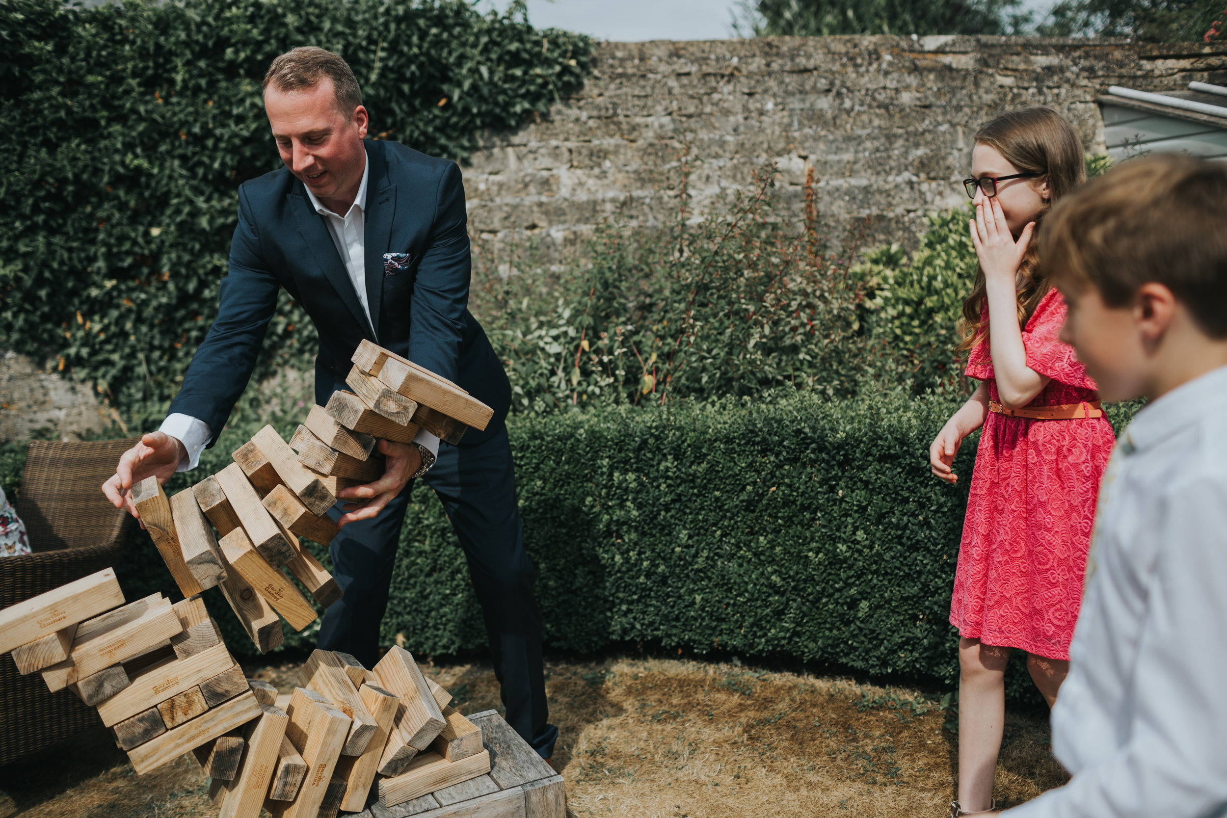 Adult fails at giant jenga at outdoor wedding, while children mock him. 