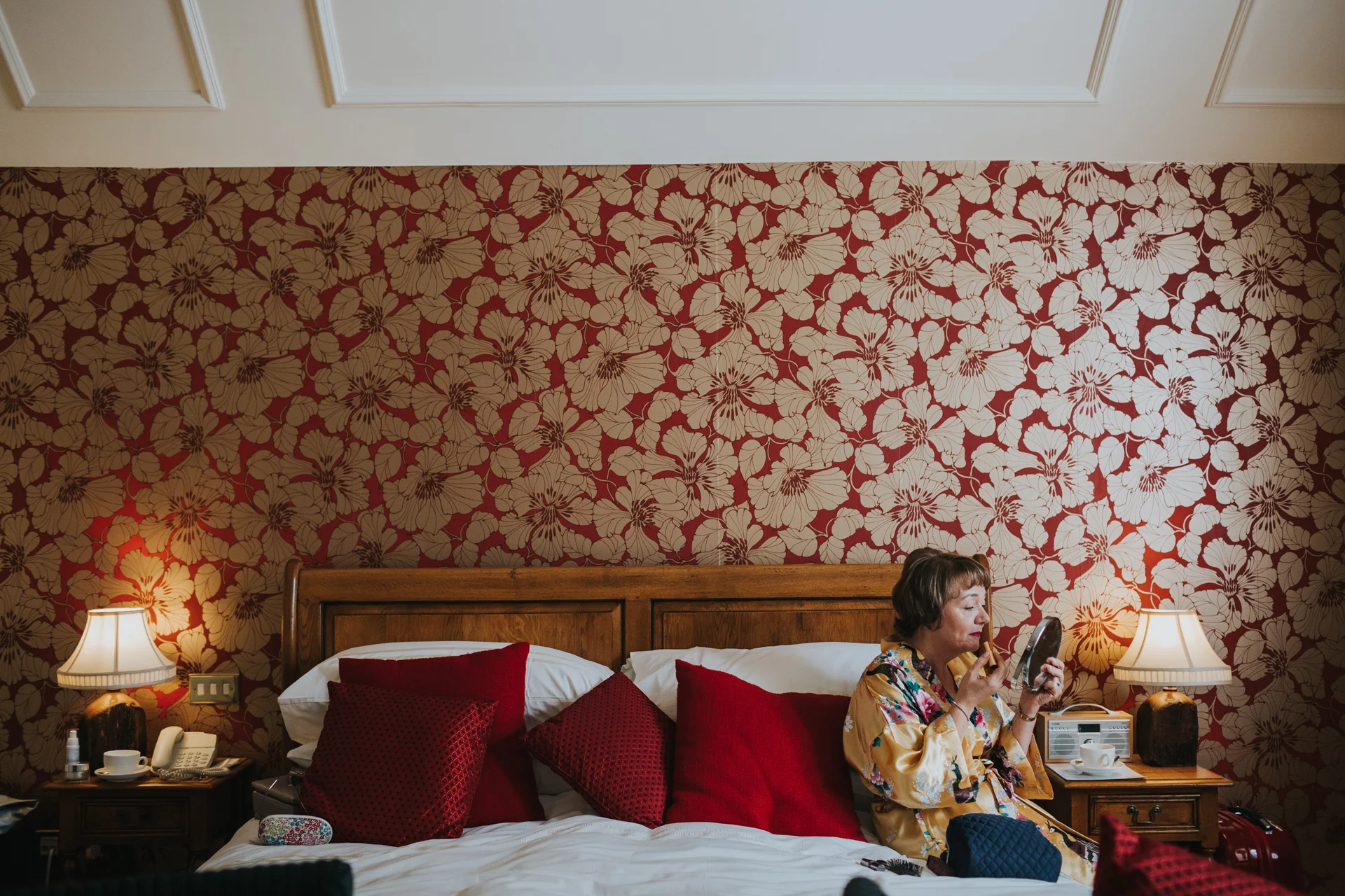 Mother of the bride sits on a bed putting on her lipstick in front of a wall of very loud red and gold floral wall paper framed by two matching lamps. 