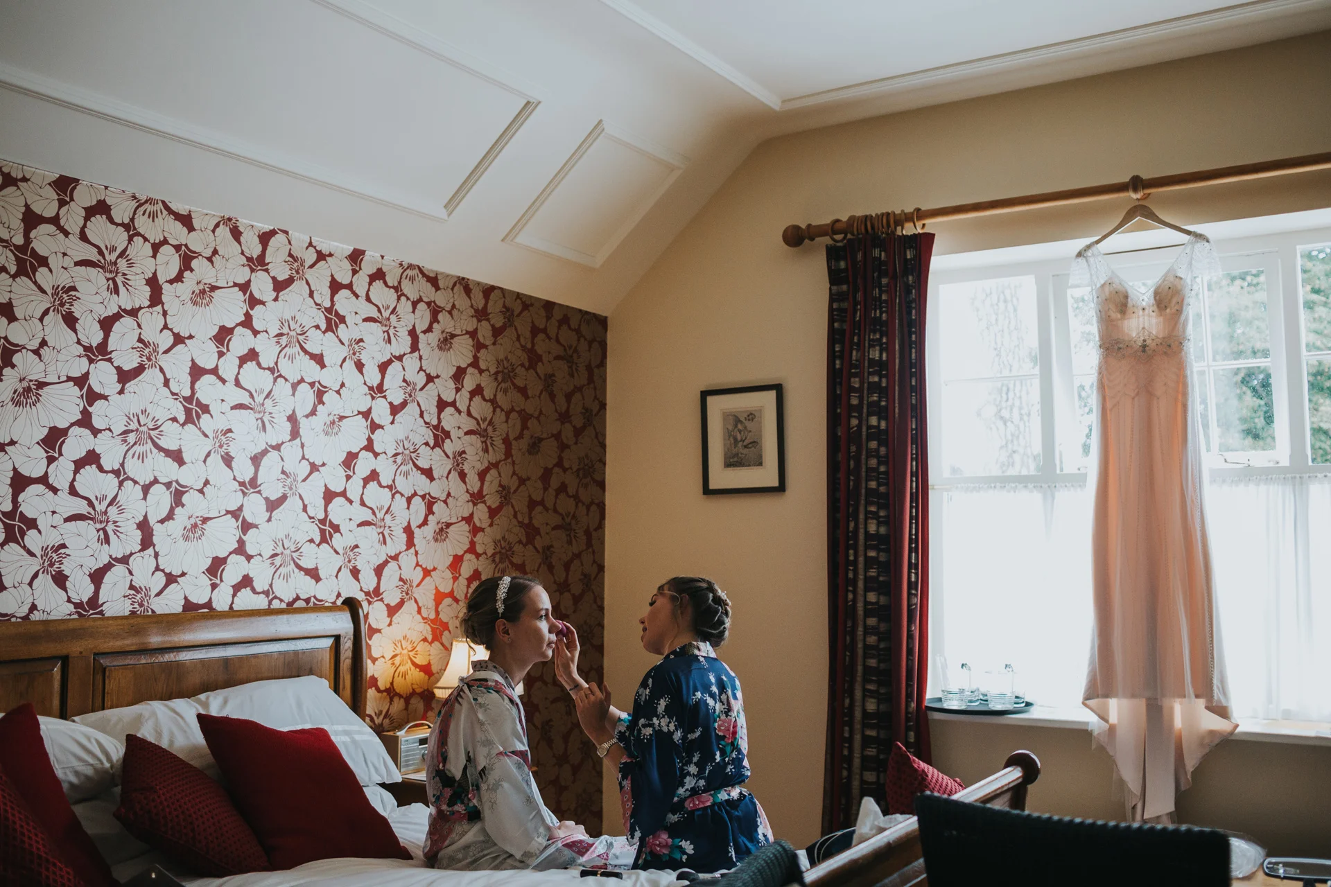 Bride has her make up done by one of her bridesmaids as her wedding dress hangs in the window at Goldstone Hall, Market Drayton. 