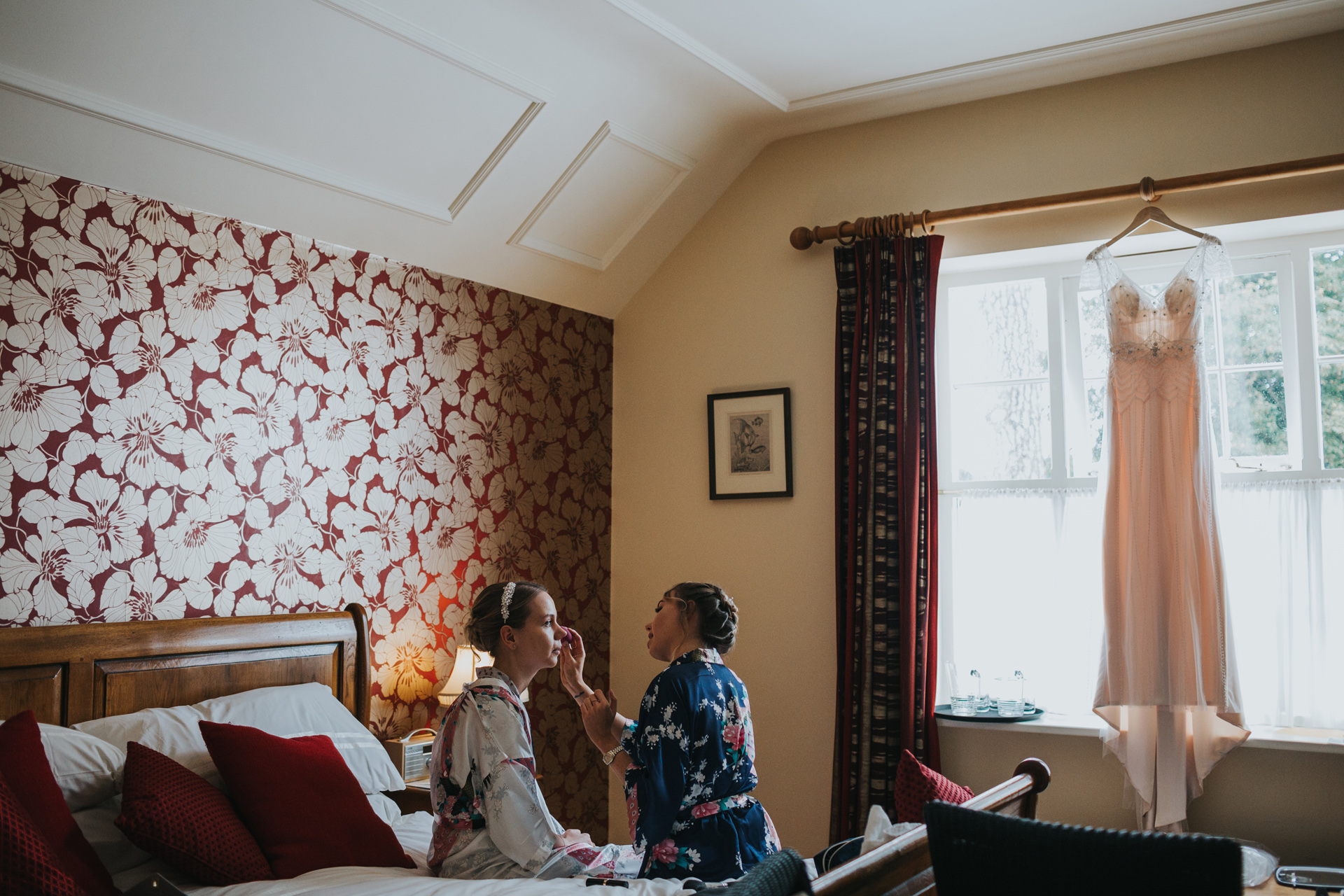 Bride has her make up done by one of her bridesmaids as her wedding dress hangs in the window at Goldstone Hall, Market Drayton.