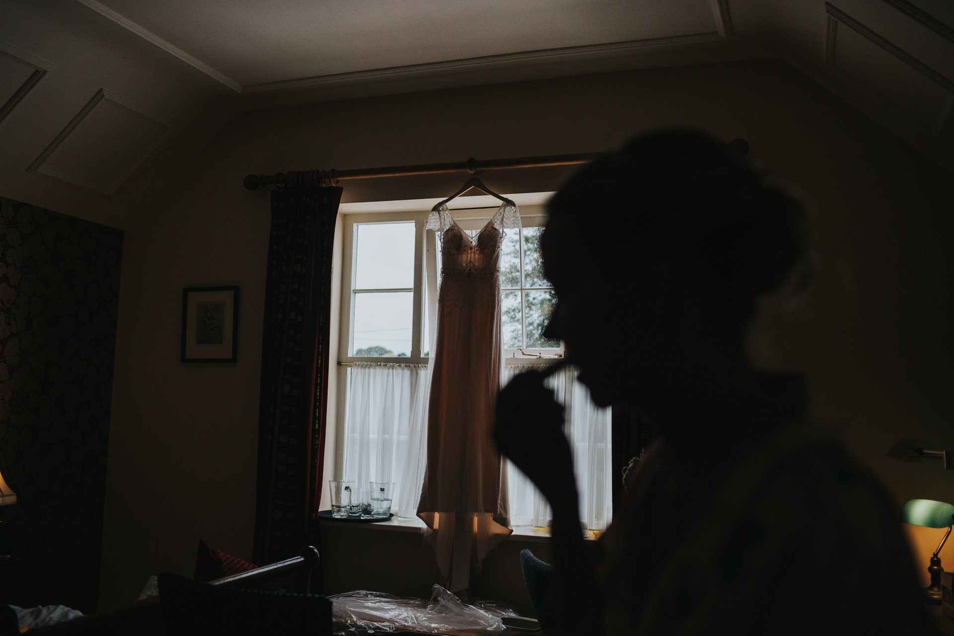 Silhouette of bride putting on her lipstick as her wedding dress hangs in the window behind her. 