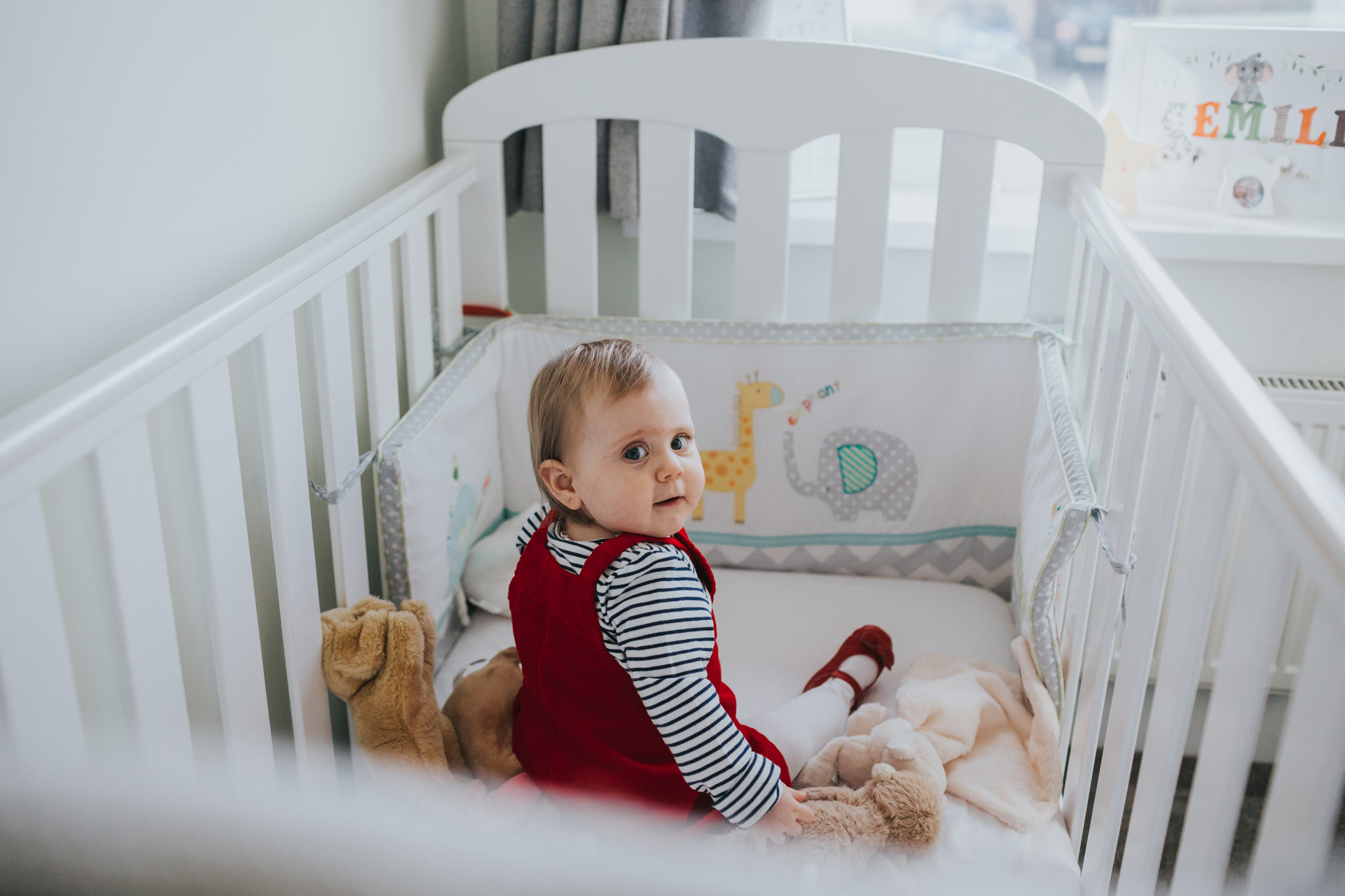 Child looks back at the camera as she sits in her cot with her cuddly toys.